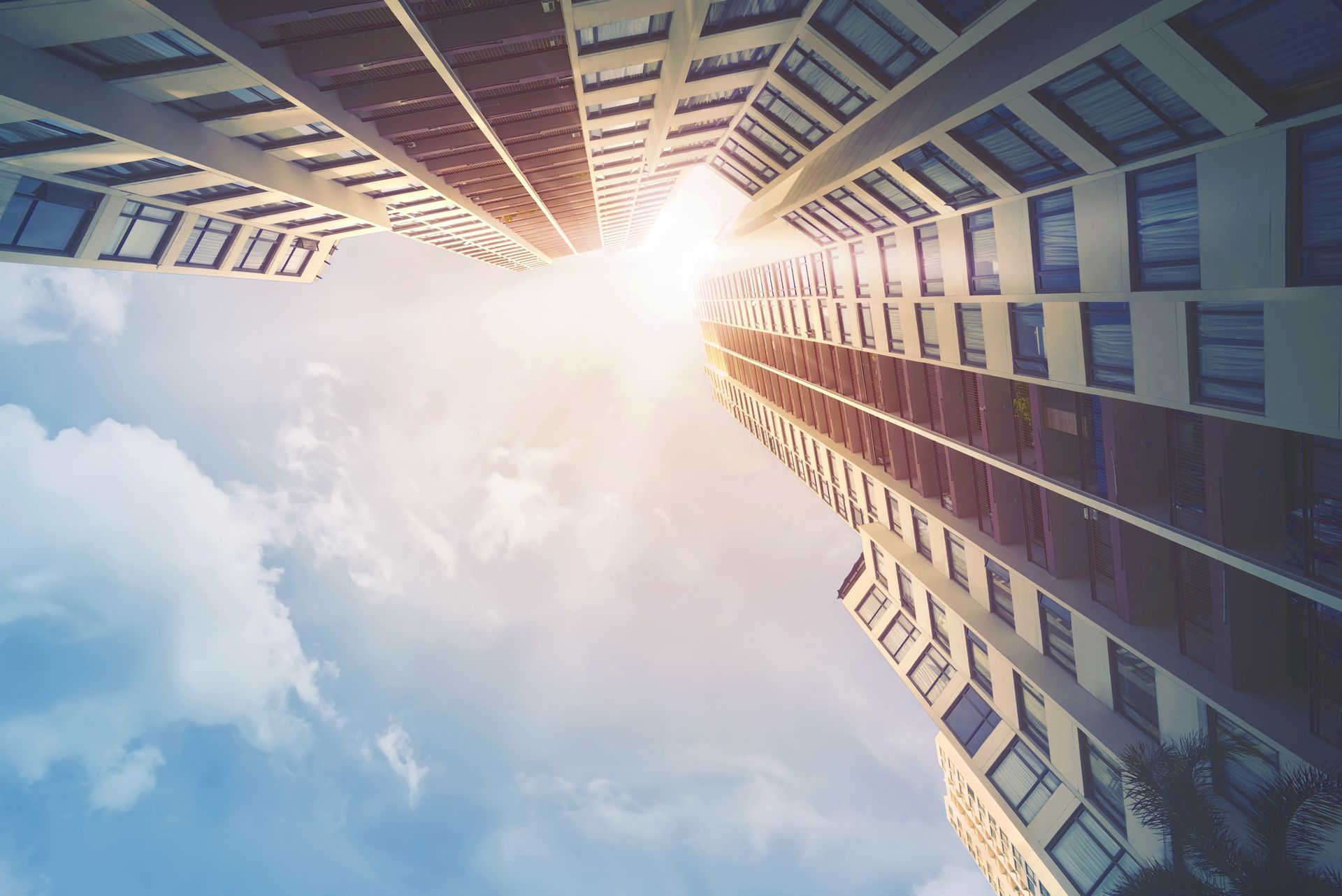 Looking up between tall buildings toward a bright sun and blue sky with clouds