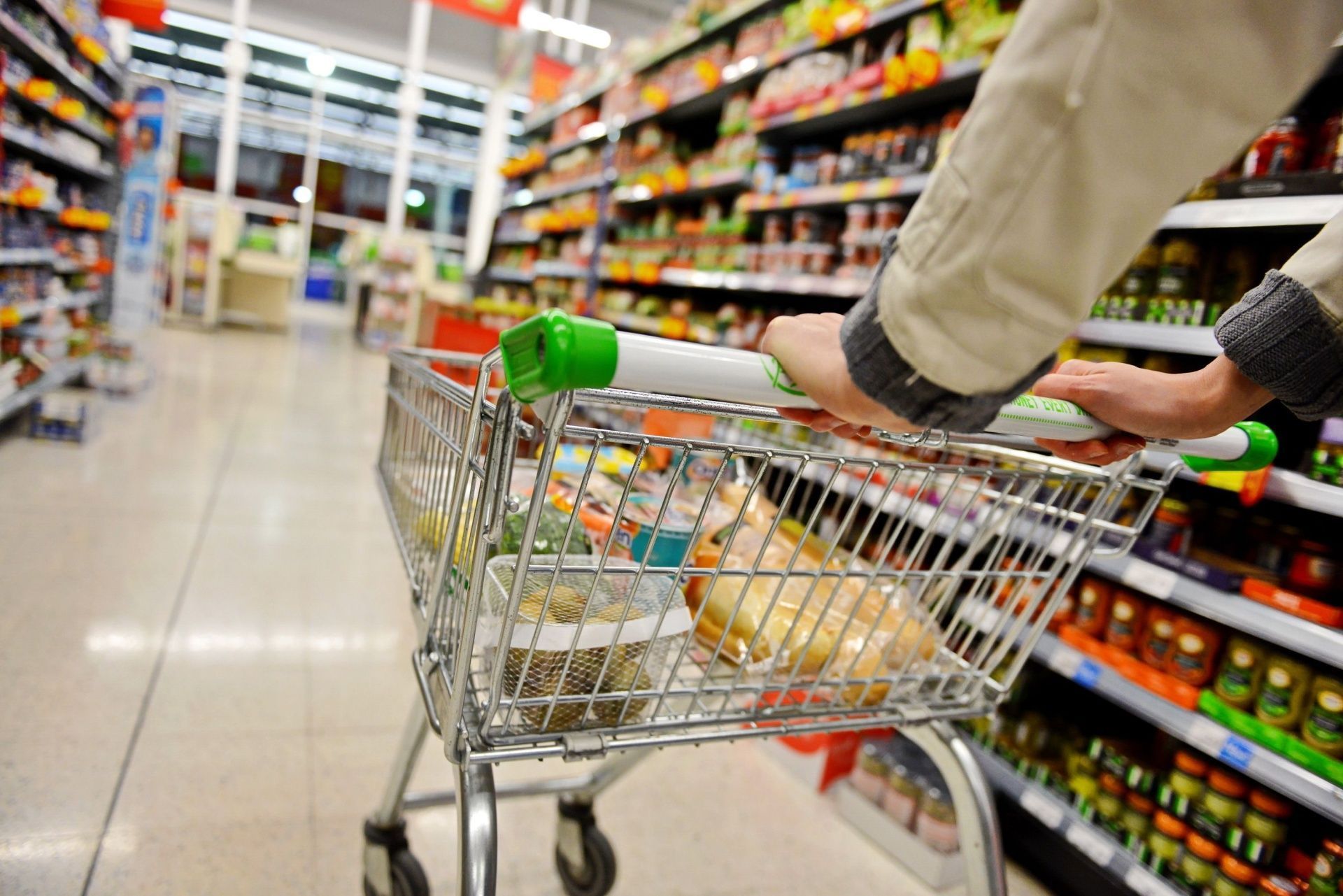 Person pushing a shopping cart down a grocery store aisle, with shelves stocked on both sides.