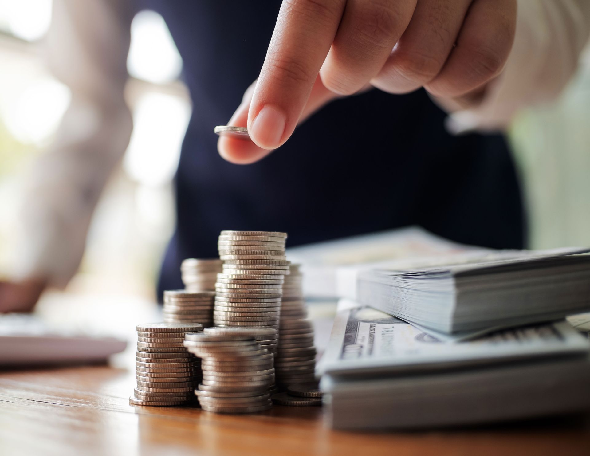 Hand placing a coin on stacked coins beside bundles of cash on a table