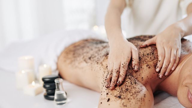 Spa massage with a client covered in coffee scrub, hands massaging the back beside candles and bottles.