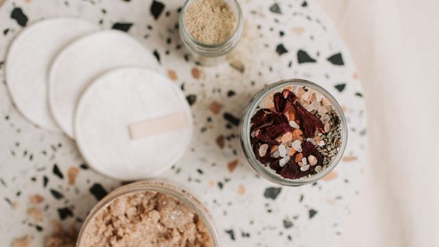 Flat lay of skincare jars, white cotton pads, and a glass bowl of dried flower bath salts on terrazzo surface