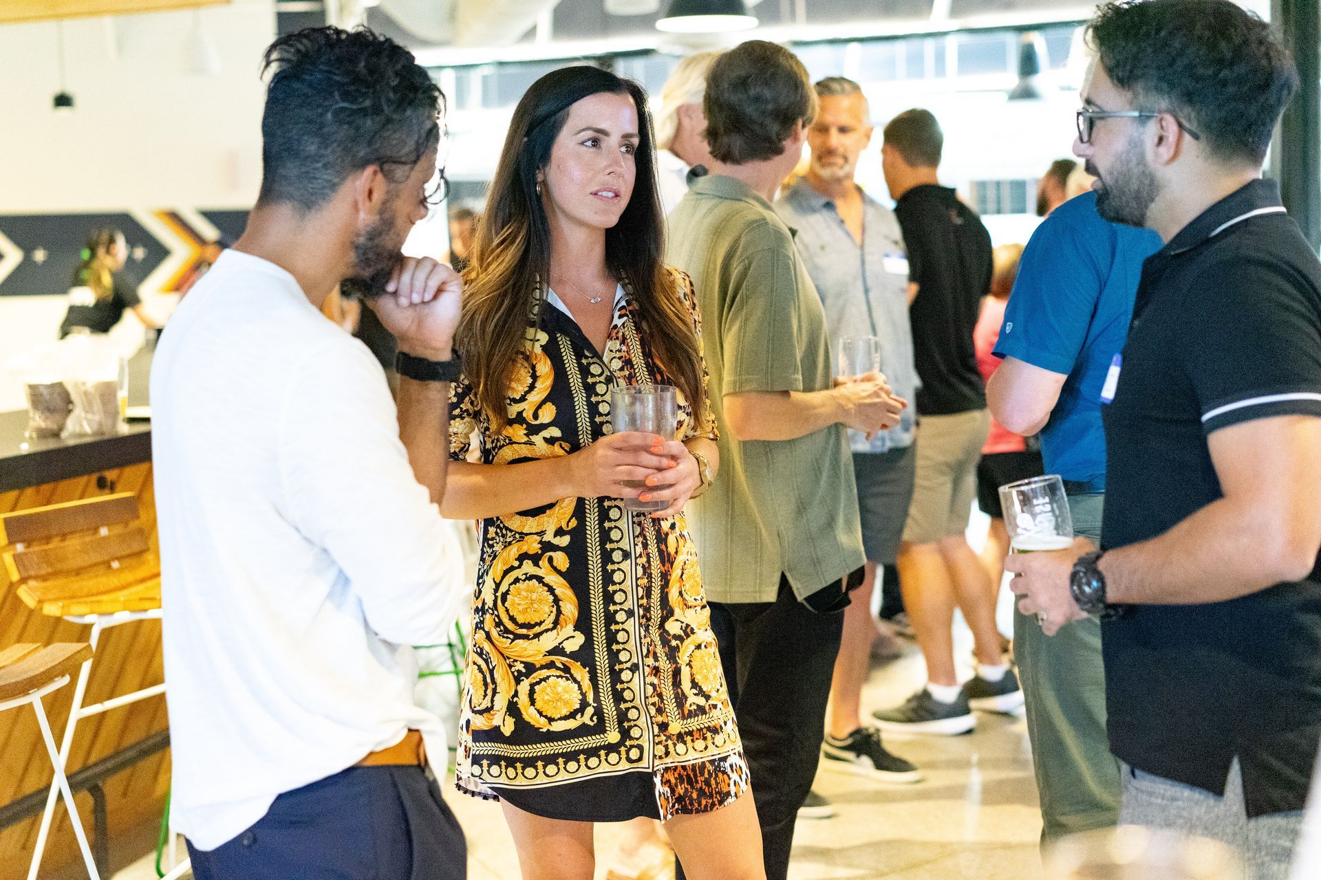 People chatting at a social gathering indoors, with drinks in hand and a bright, modern setting.