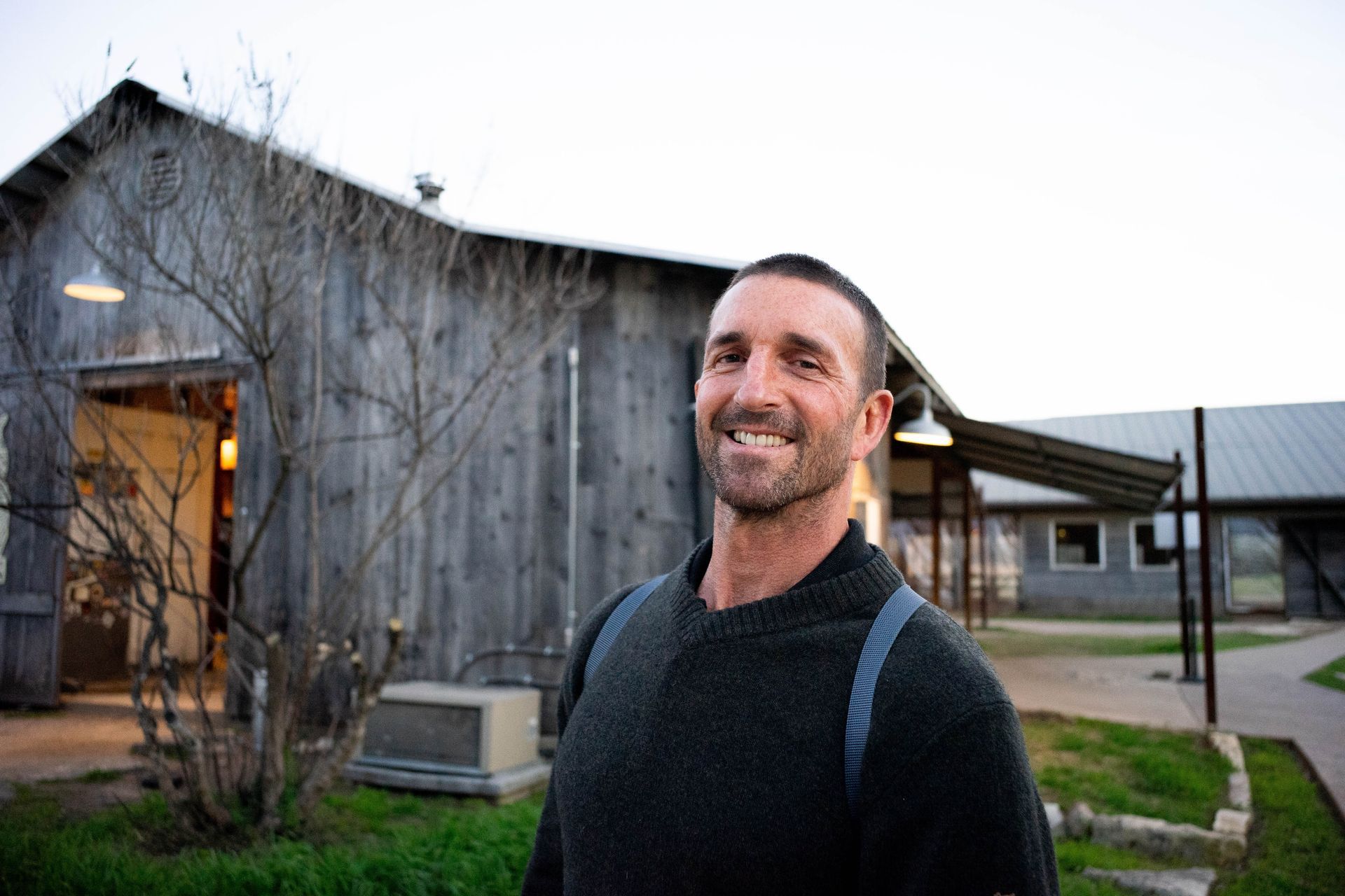 Smiling man standing outside a rustic barn at dusk, with lit windows and a grassy yard.