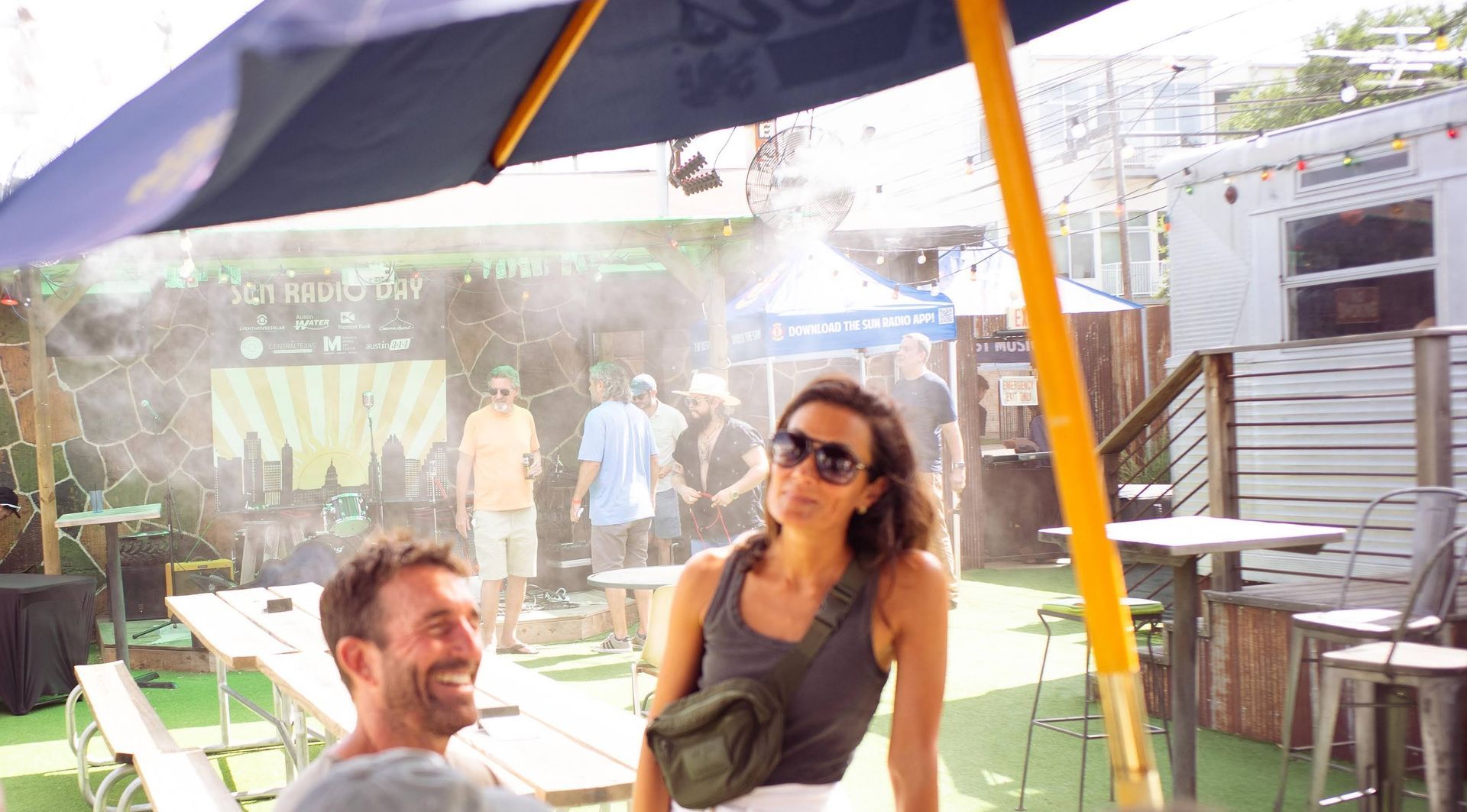 Two people laughing under a patio umbrella at an outdoor café in bright sunlight