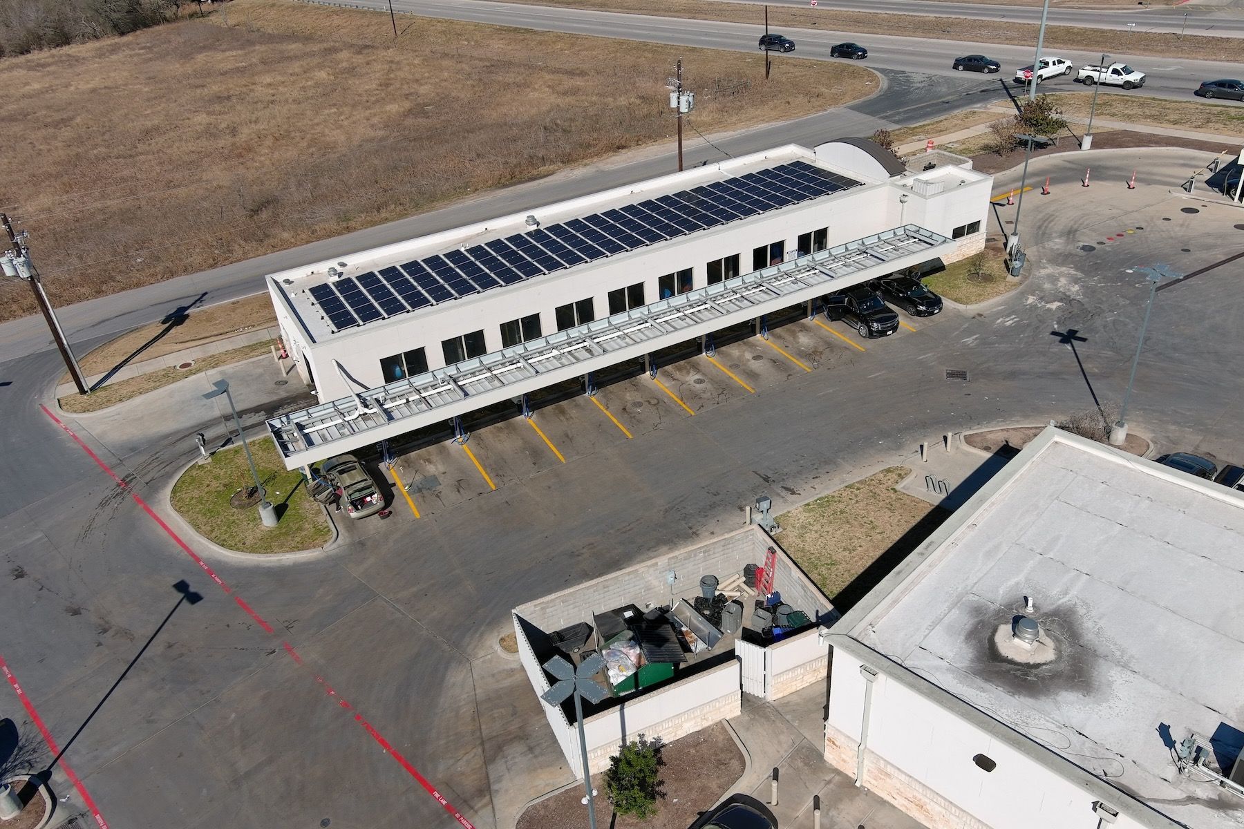 Aerial view of a white building with solar panels on the roof in a parking lot, near an open lot and another building
