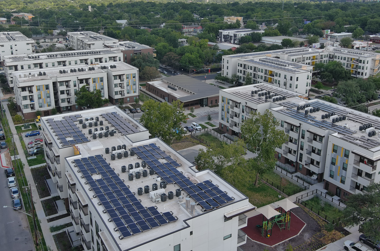 Aerial view of modern apartment buildings with rooftop solar panels and surrounding green trees.