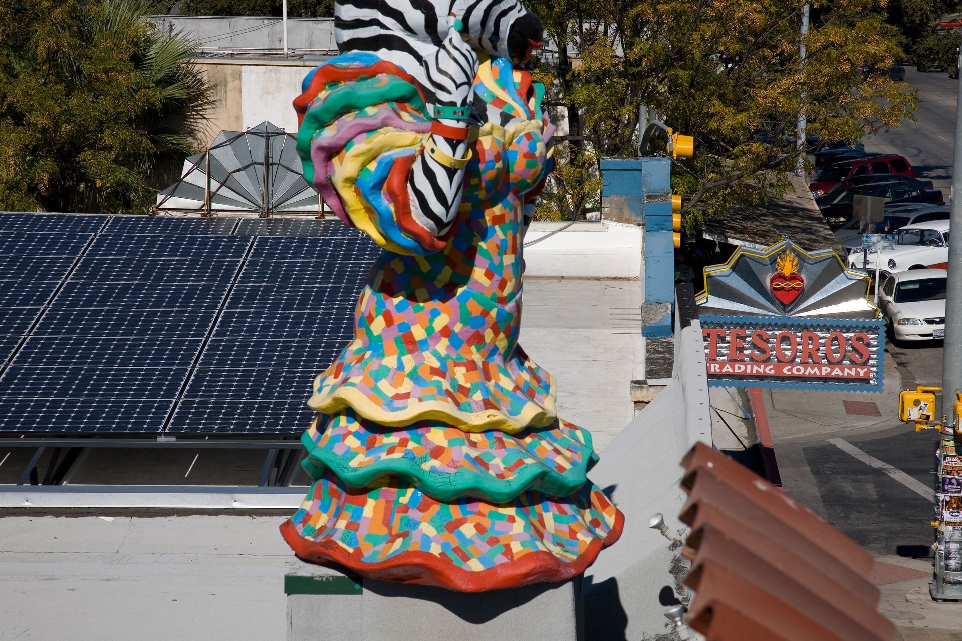 Colorful mosaic sculpture on a rooftop beside solar panels, with a sign and parked cars in the background.