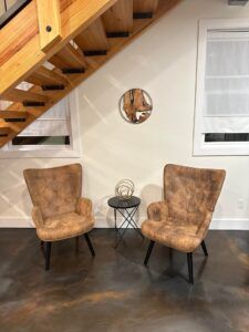 Two tan armchairs face each other, with a small table between them under a wooden staircase.