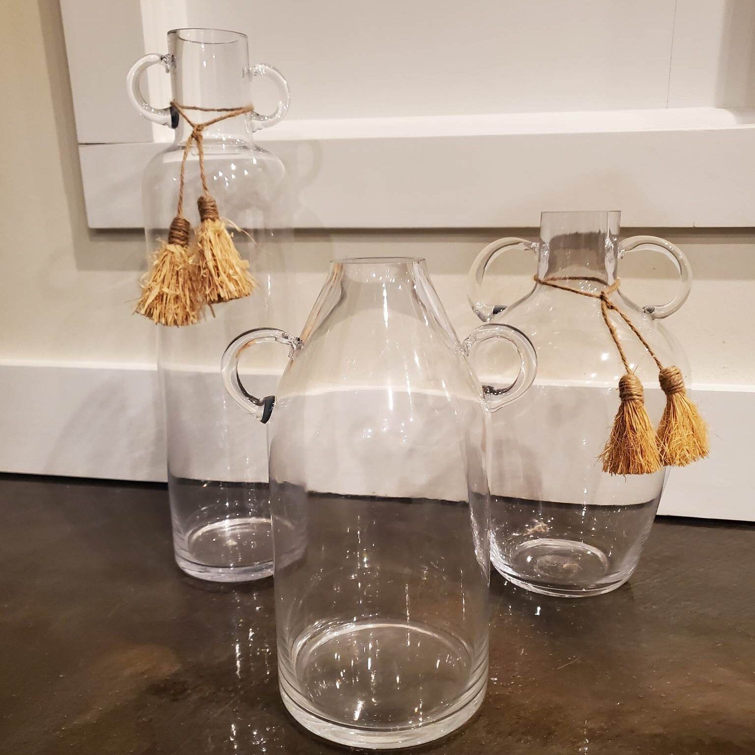 Three clear glass vases with handles and decorative tassels.