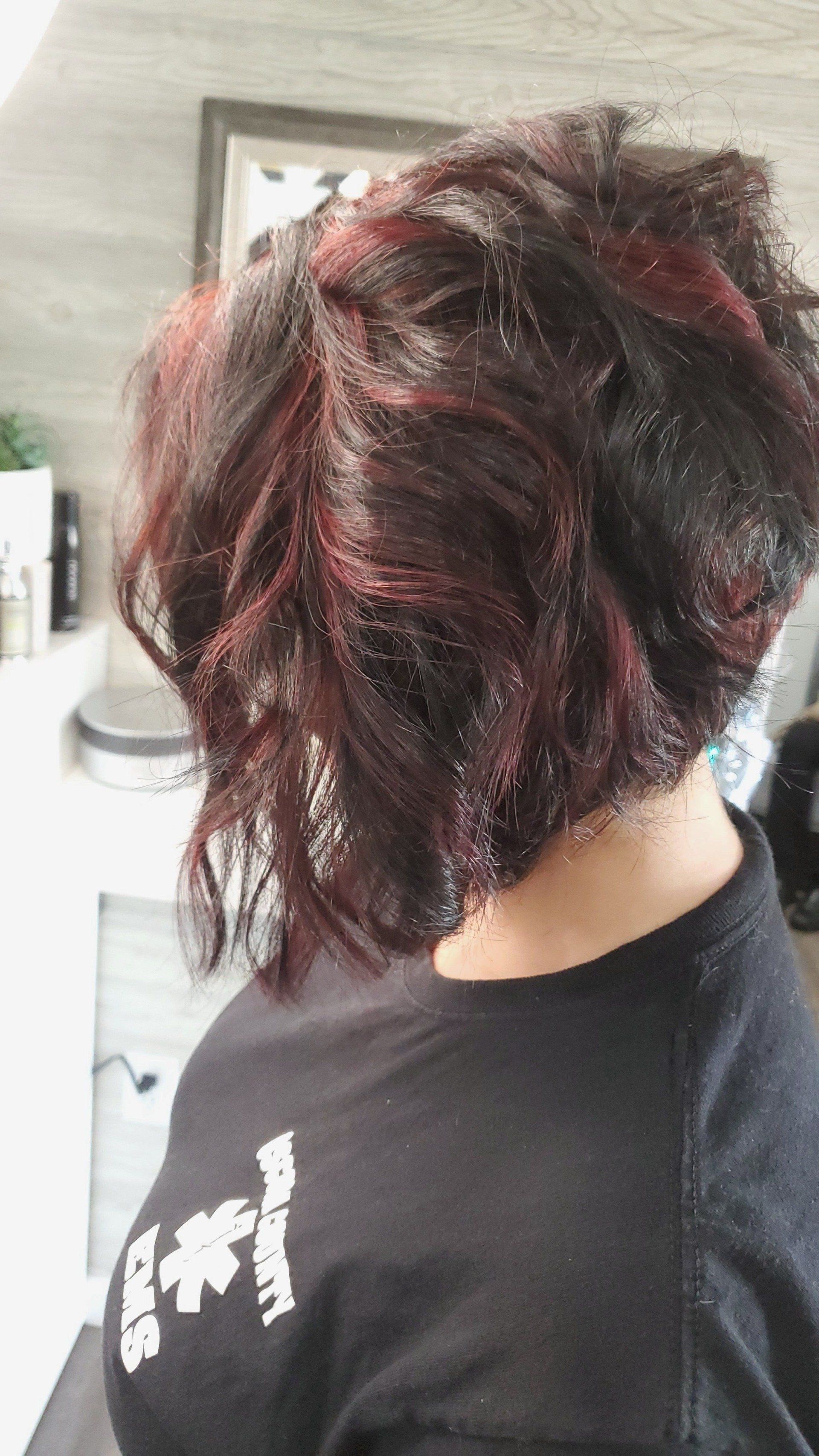 Person with layered, dark hair with red highlights. They are in a salon, wearing a black shirt.