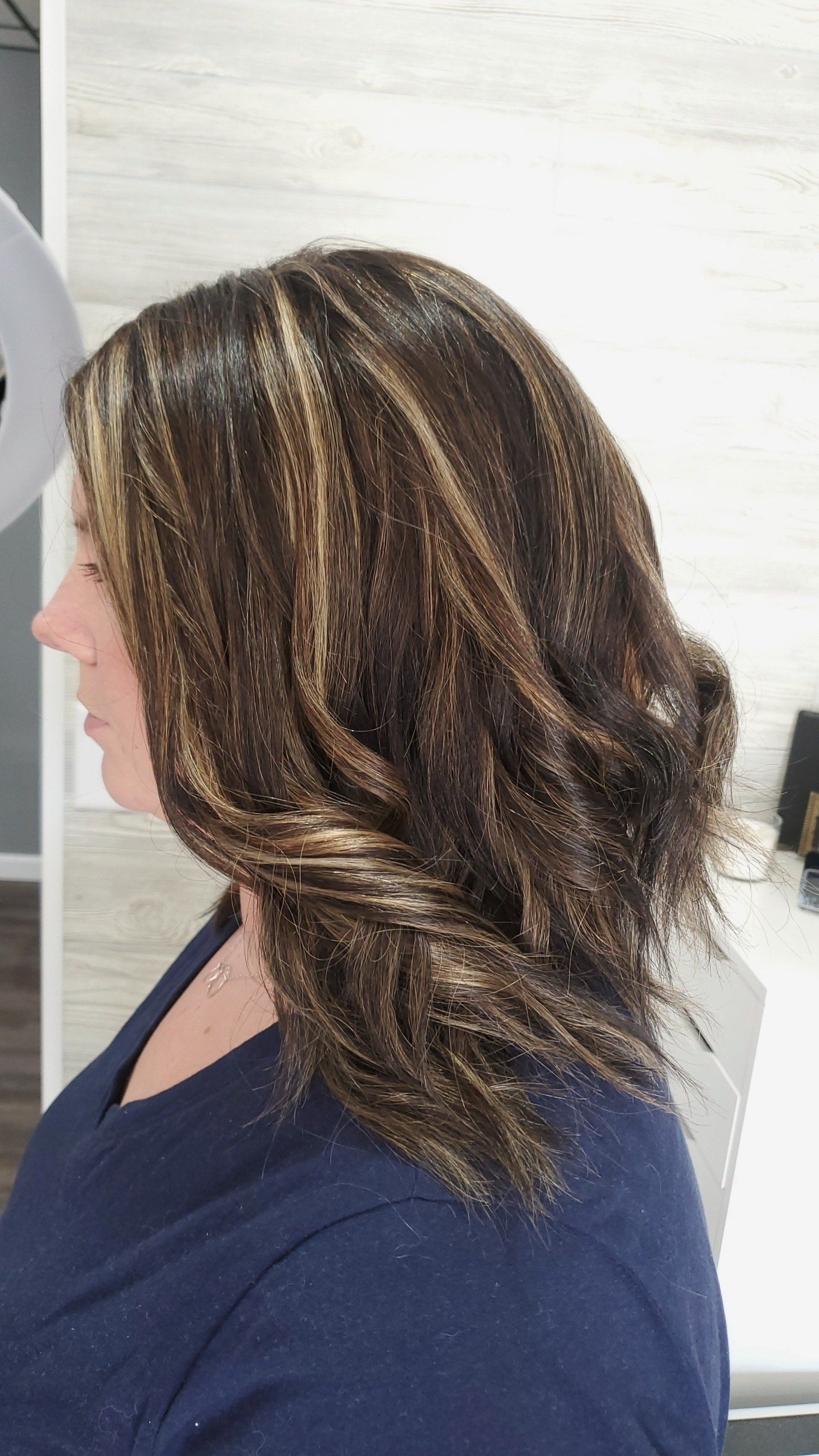 Woman with dark brown hair, highlighted with blonde. She wears a dark blue shirt and is in a salon.