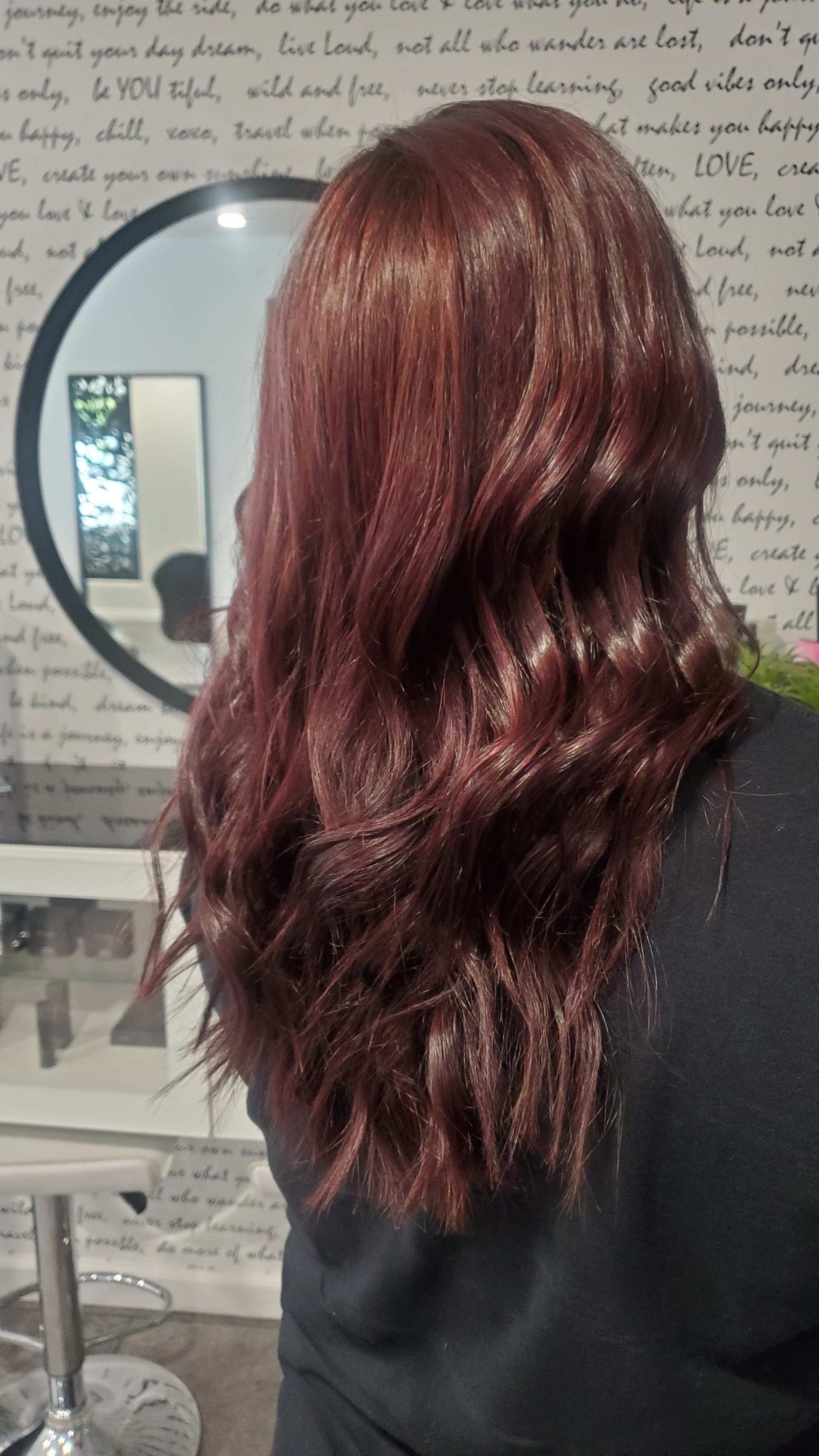 Woman with long, wavy auburn hair. Standing in front of a mirror with a decorative background, likely in a salon.