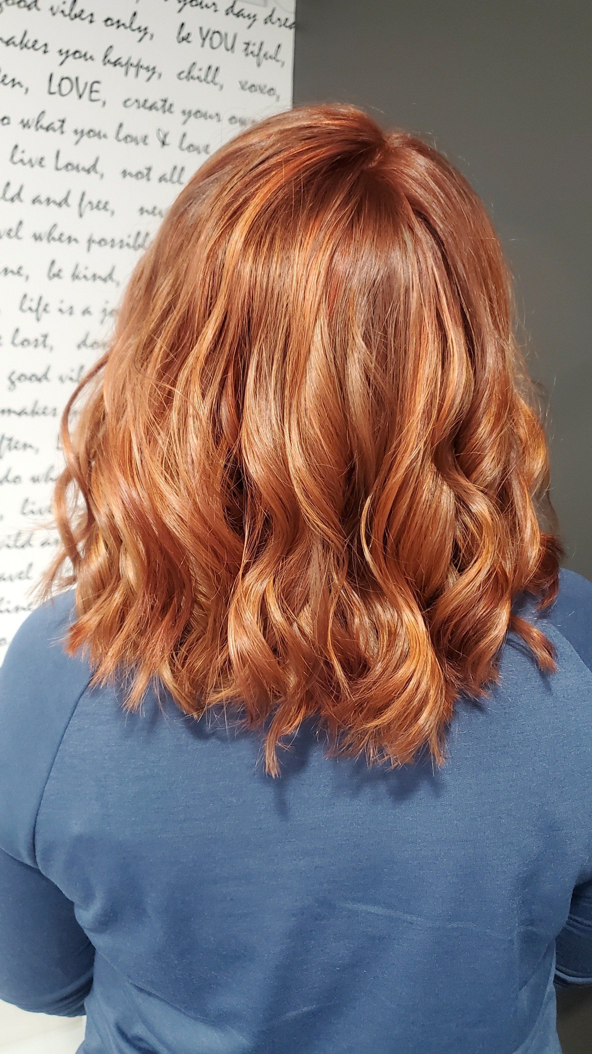 Woman with auburn wavy hair, wearing a blue shirt, stands against a white wall with inspirational text.