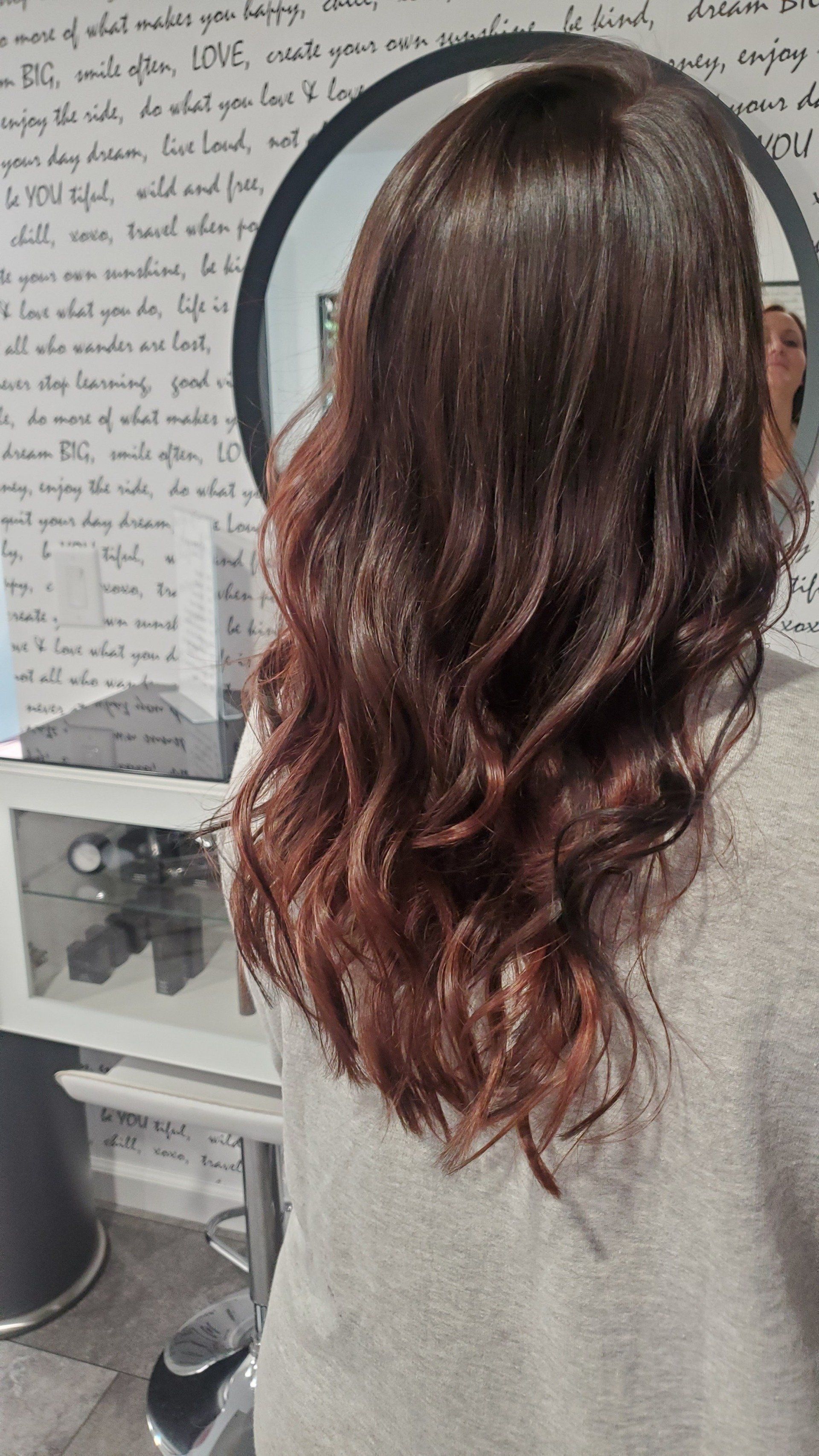 Woman with brown hair, styled with loose waves, leaning toward a circular mirror.