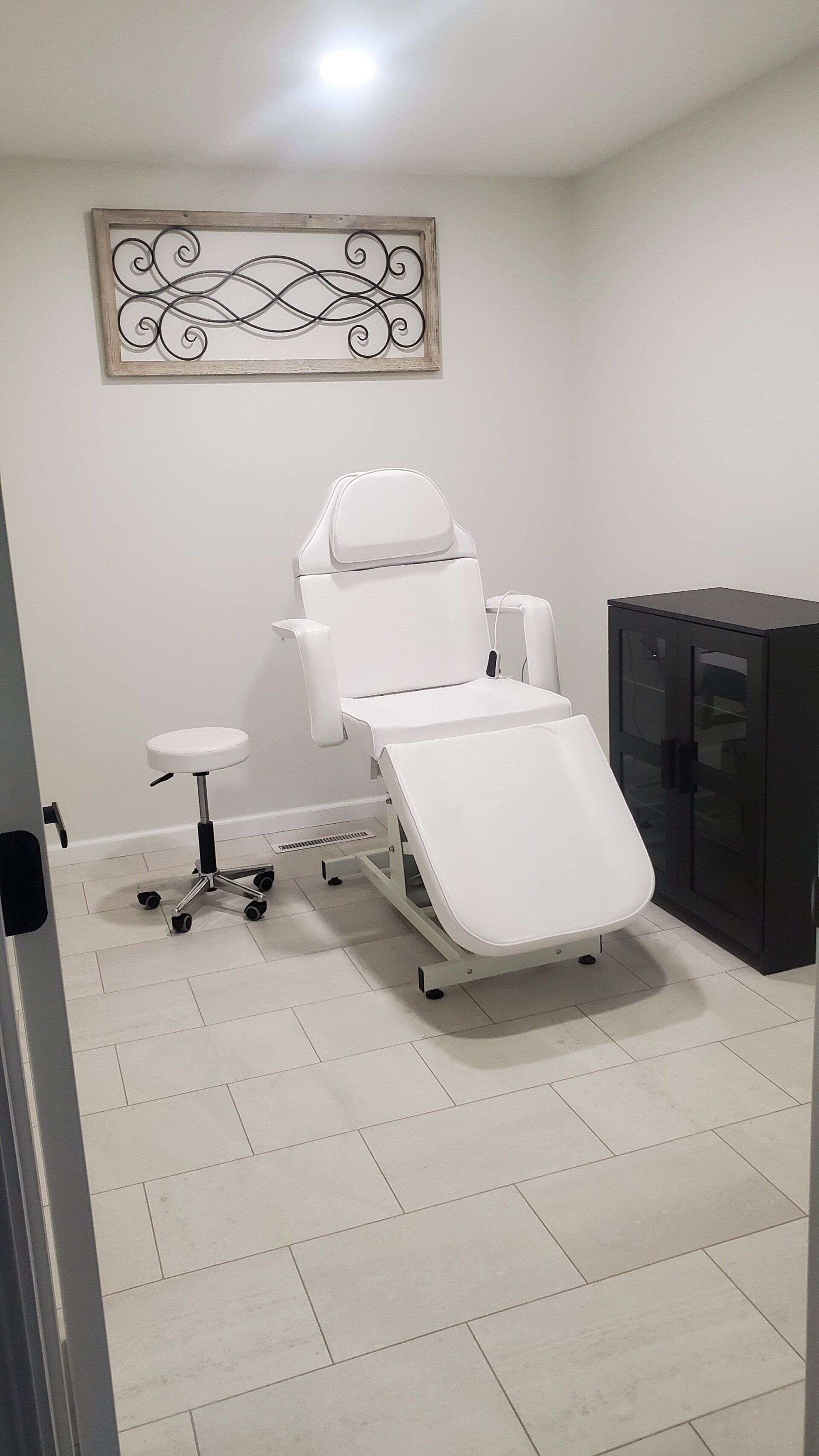 Treatment room with white chair, stool, and cabinet. Decorative wall art. Light grey tiled floor.