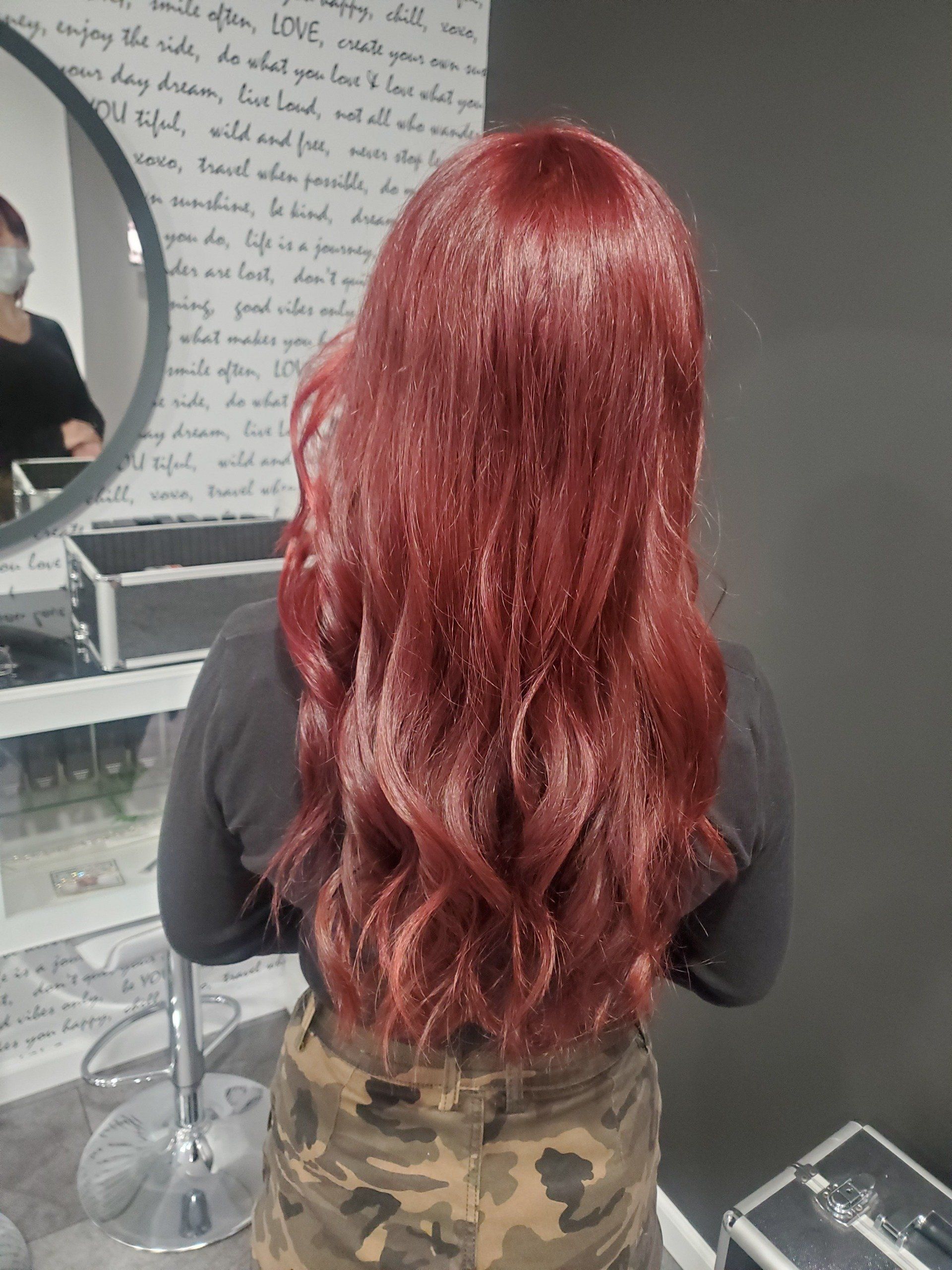 Woman with long, wavy, reddish-brown hair stands in a salon.  She wears a black shirt and camo pants.
