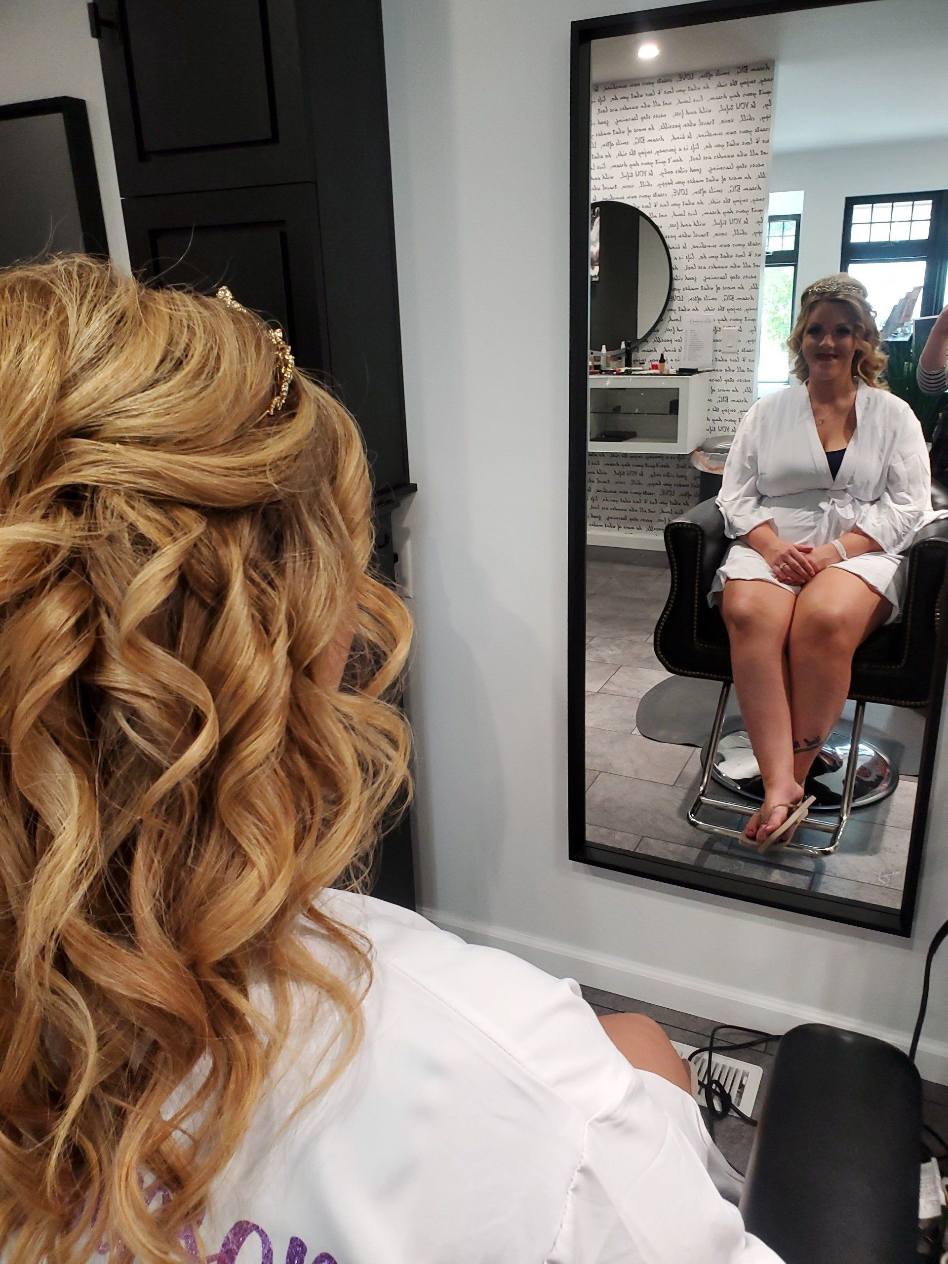 Woman with curled updo looks into a mirror at a hair salon. She wears a white robe.