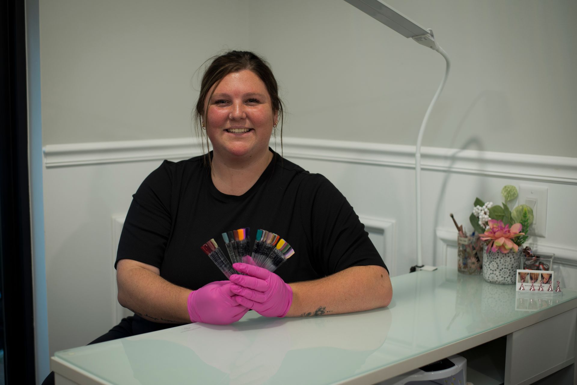Woman in pink gloves holding nail polish samples, smiling at a table with lamp, flowers, and containers.