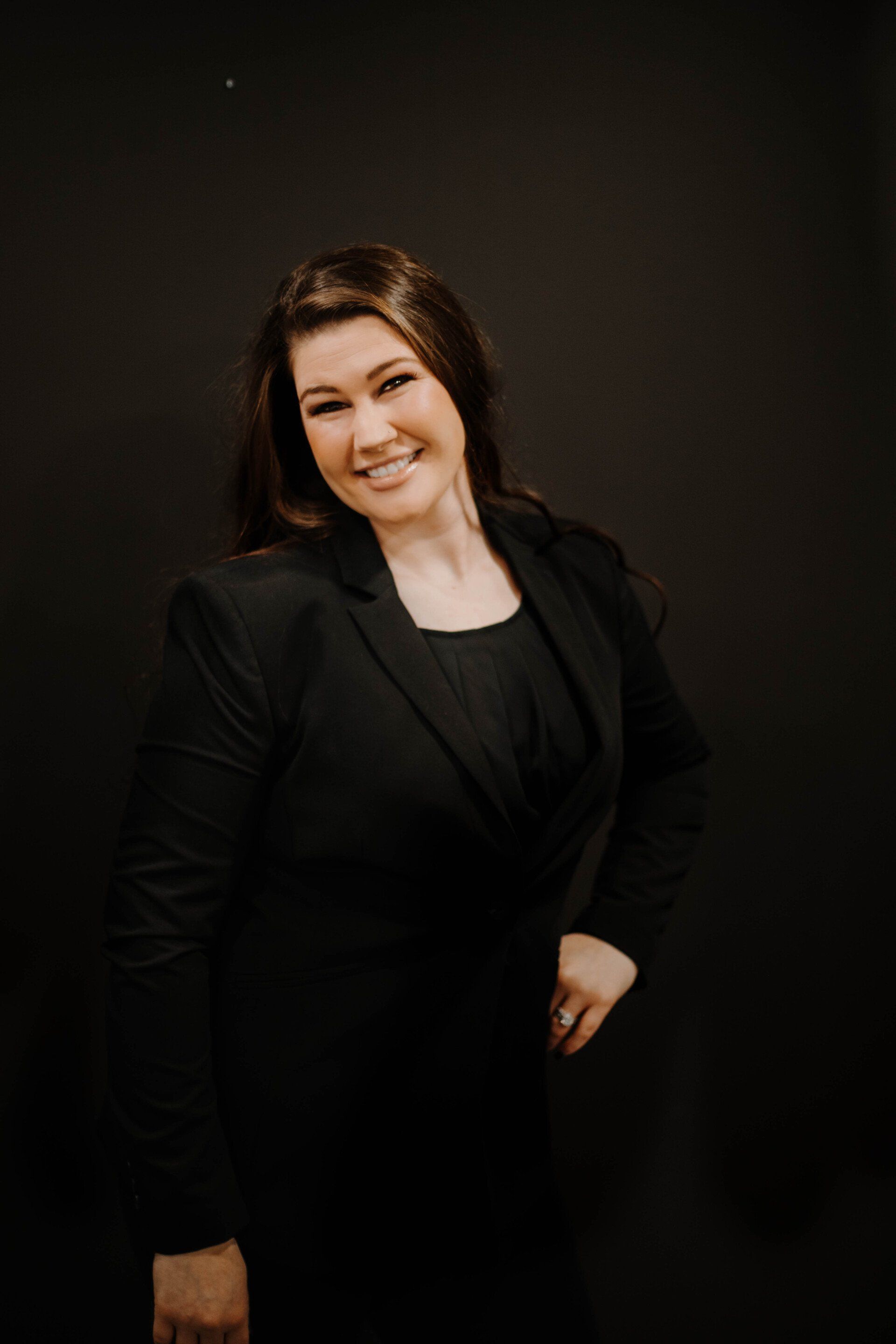 Woman in black blazer and dress smiles, standing against a dark backdrop.