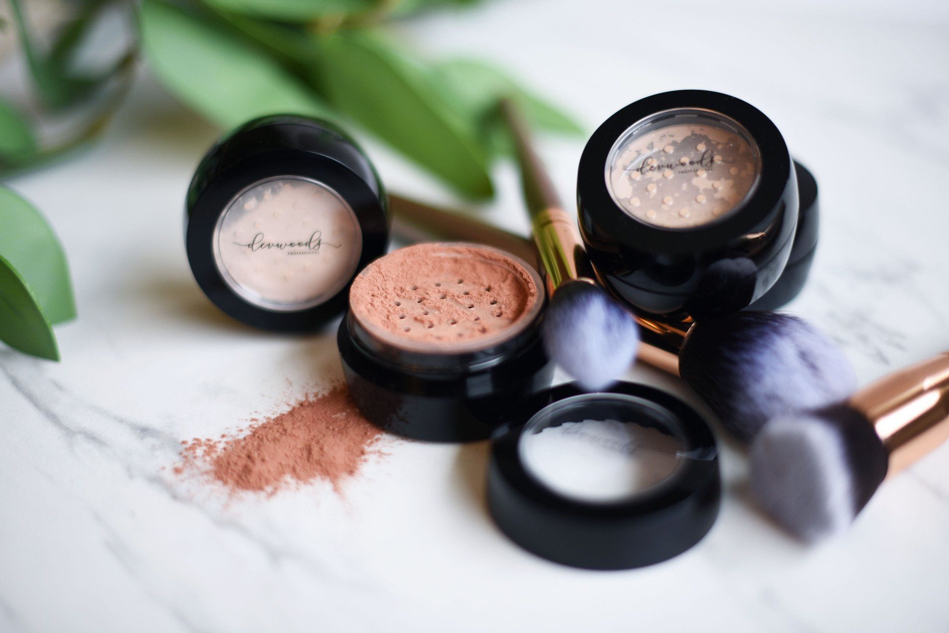 Makeup products (jars and brushes) on a marble surface, with scattered powder.