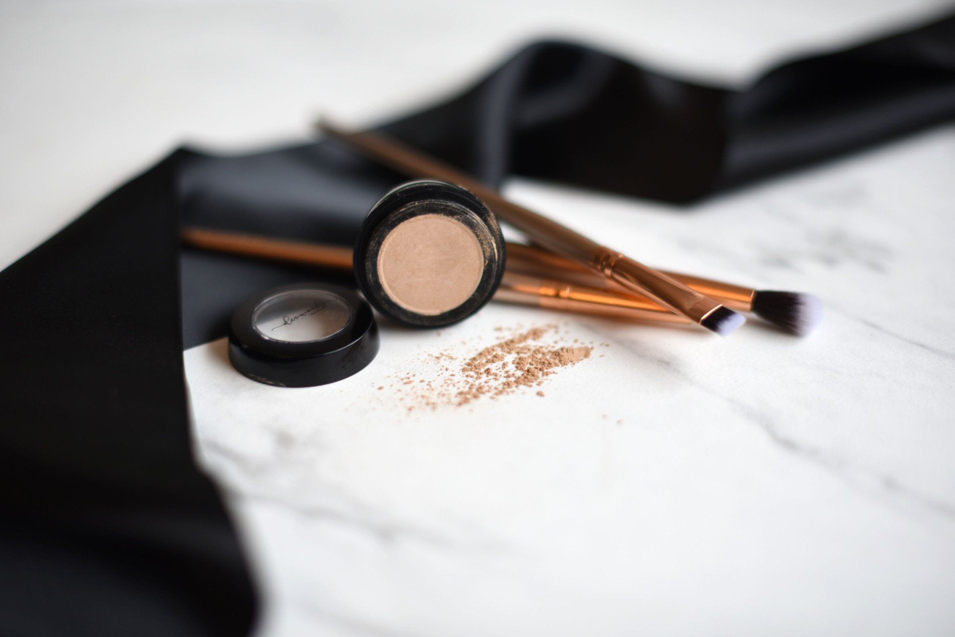 Open eyeshadow jar with spilled powder, brushes, and black ribbon on marble.