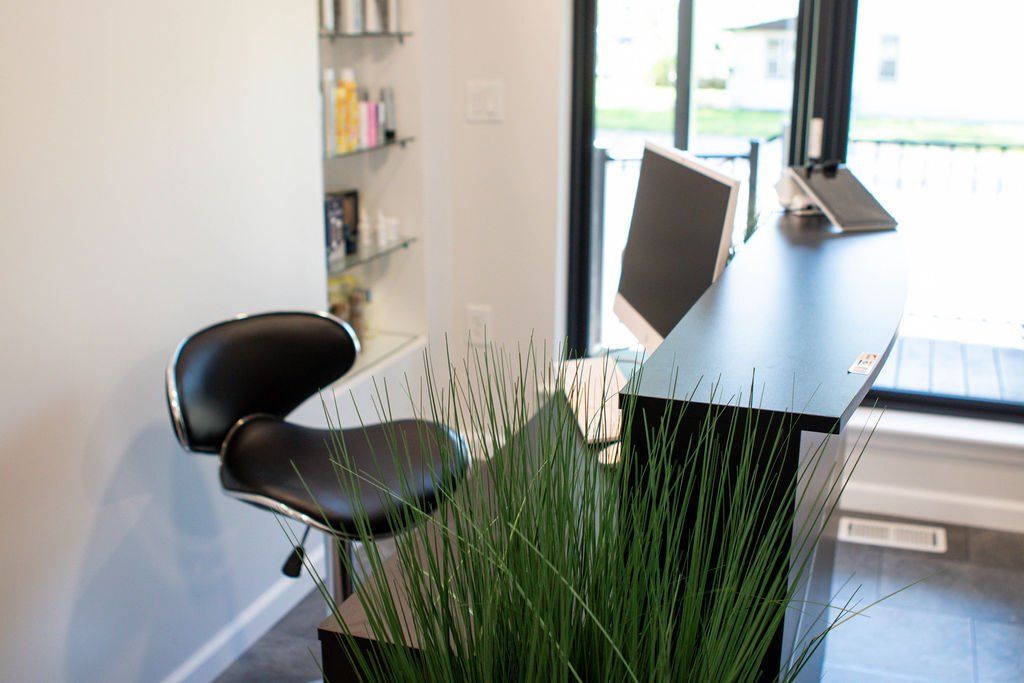 Reception desk with a black chair, monitor, and shelf with products; near a window.