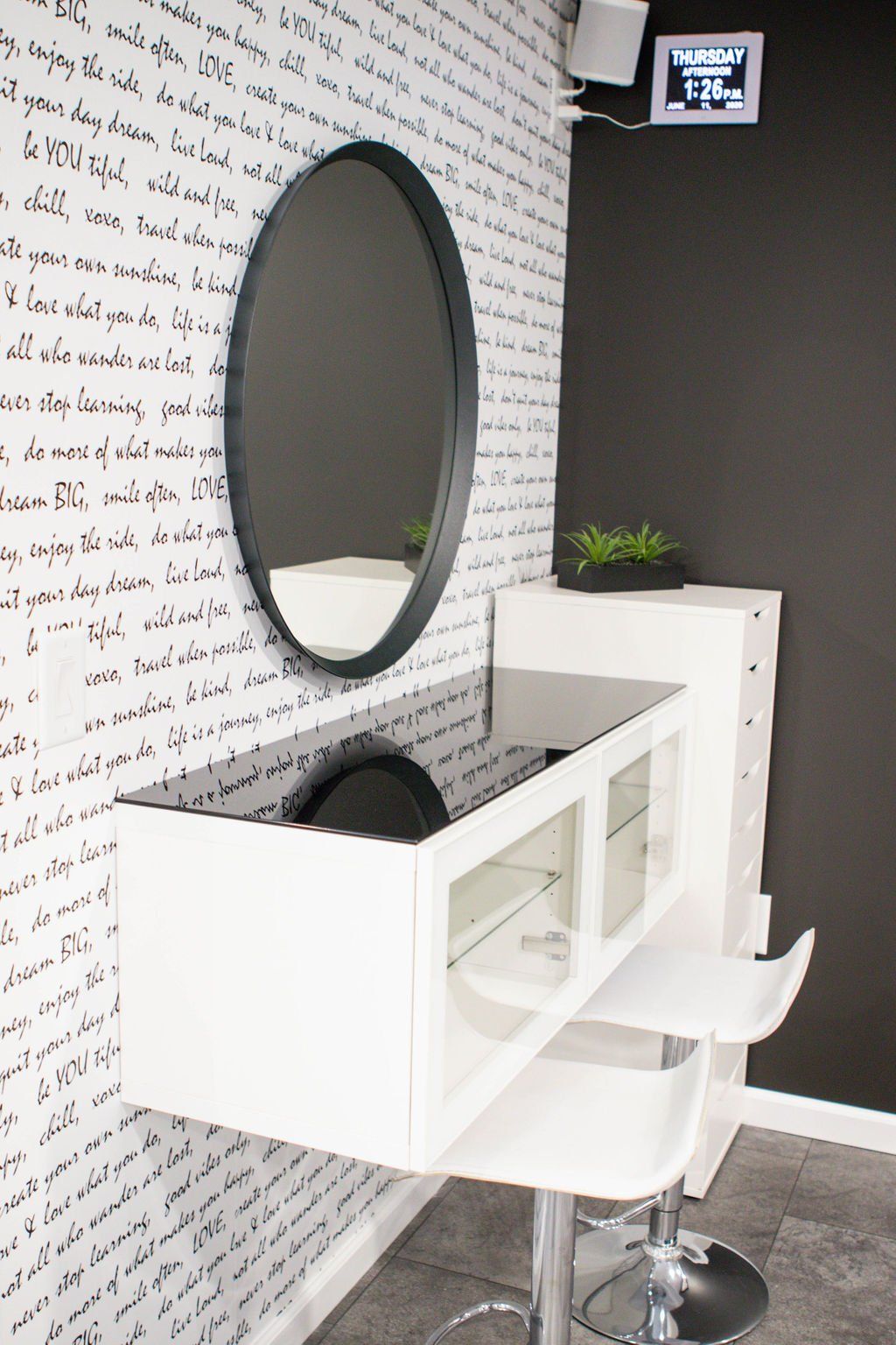 White vanity with oval mirror, black trim, and script wallpaper. Two white barstools below.