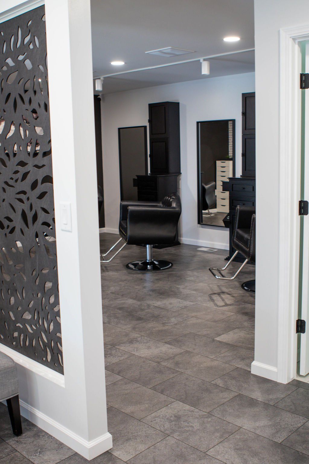 Interior view of a modern hair salon with dark grey flooring, black styling chairs, and mirrors. White walls and decorative panel.