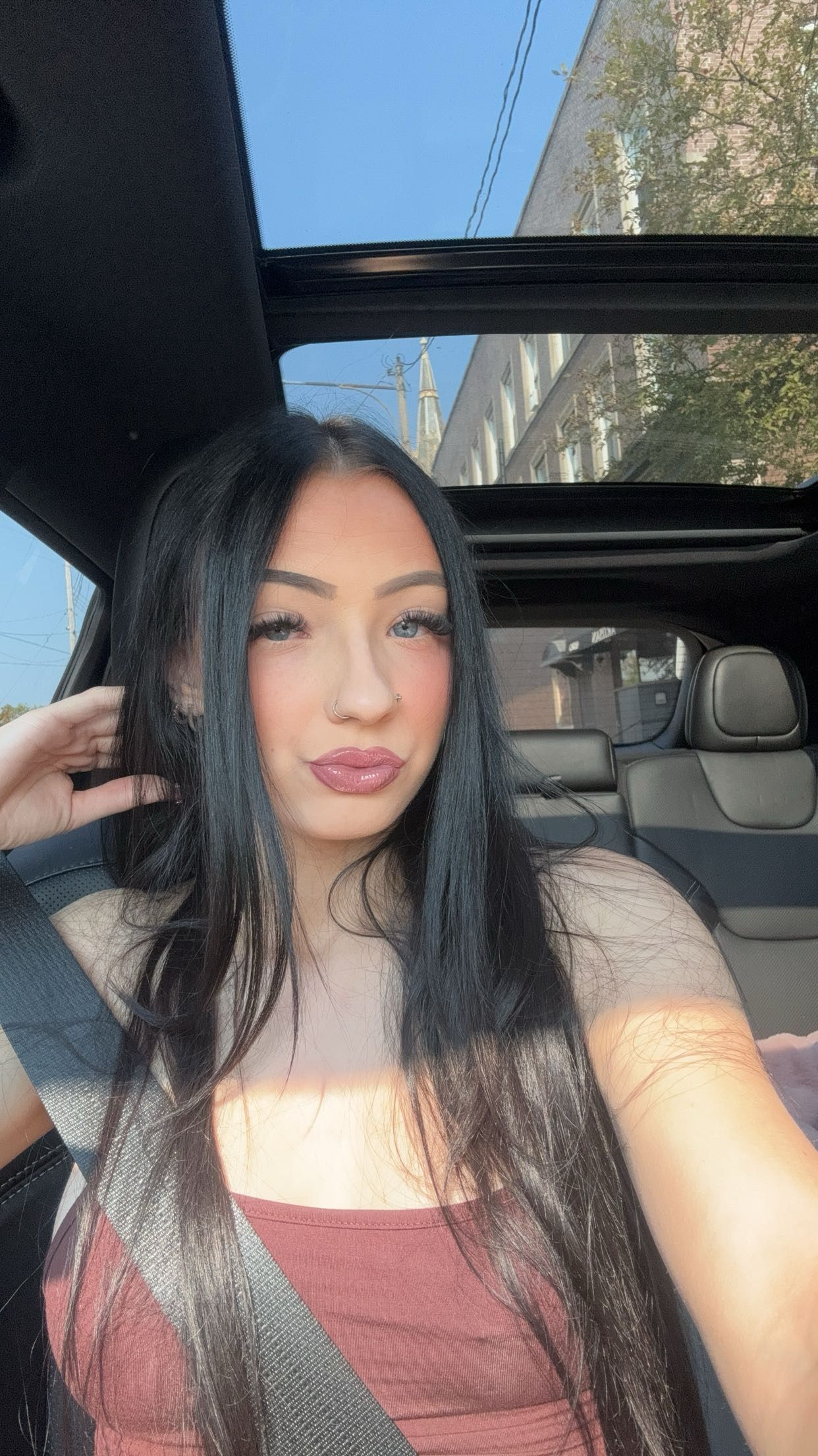 Woman with long dark hair pouting, wearing a maroon top, in a car with a sunroof open.