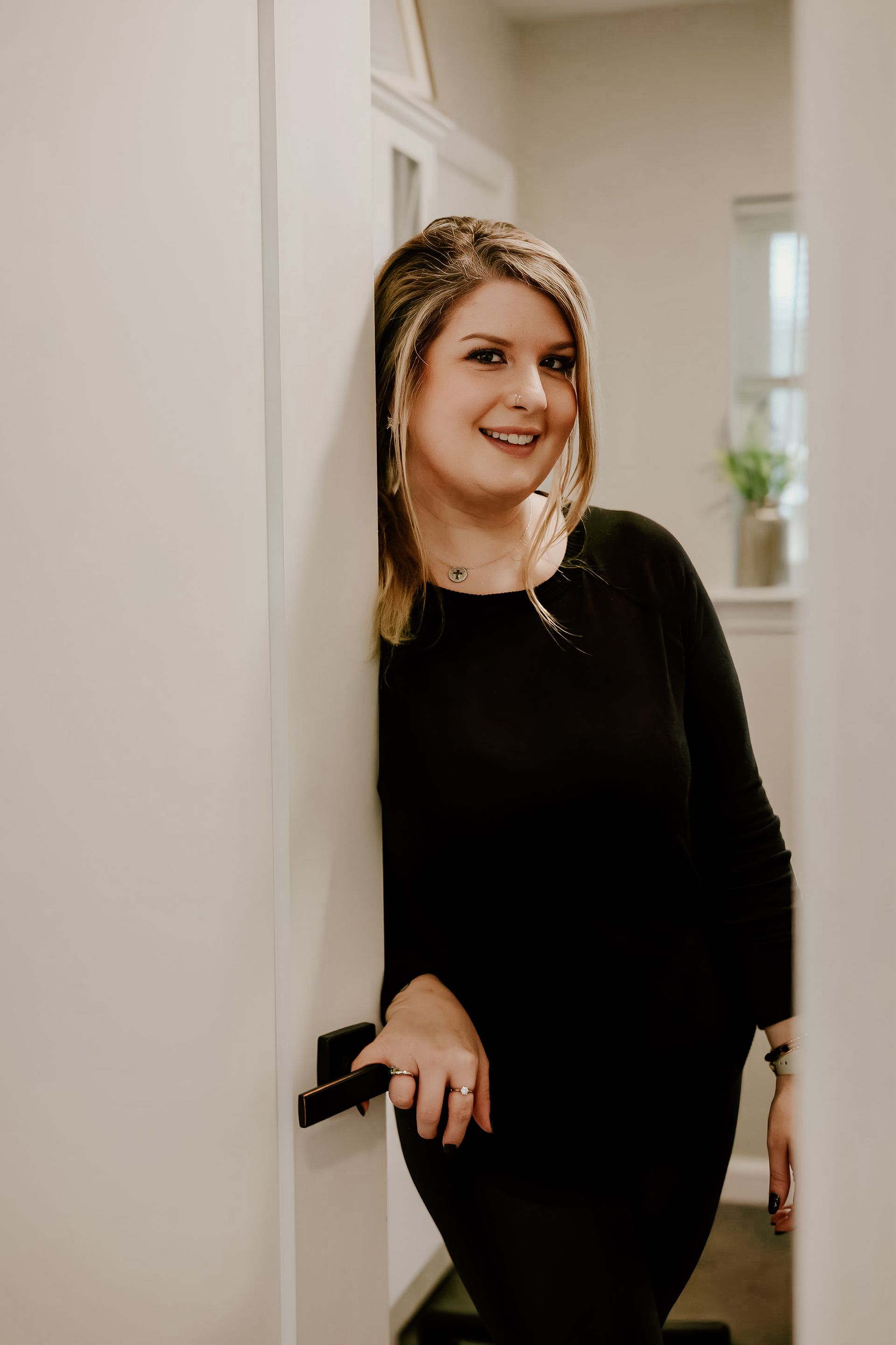 Woman in a black dress leans against a white doorway, smiling, indoors.