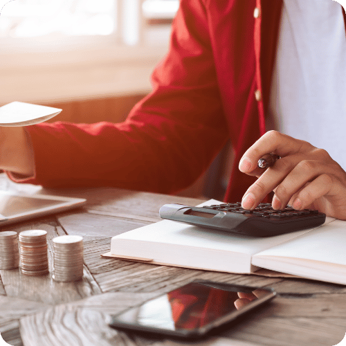 Person in red jacket using a calculator and pen on a table with a book, coins, tablet, and phone.