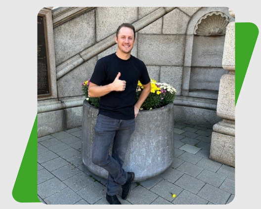 Man in black t-shirt and jeans gives thumbs up, leaning on a stone planter with flowers.