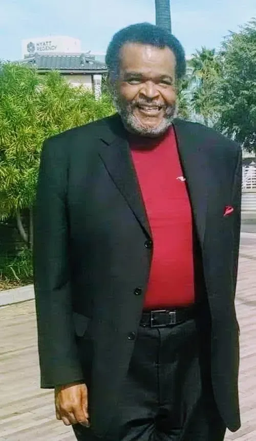 Man in black suit and red shirt smiling outdoors.