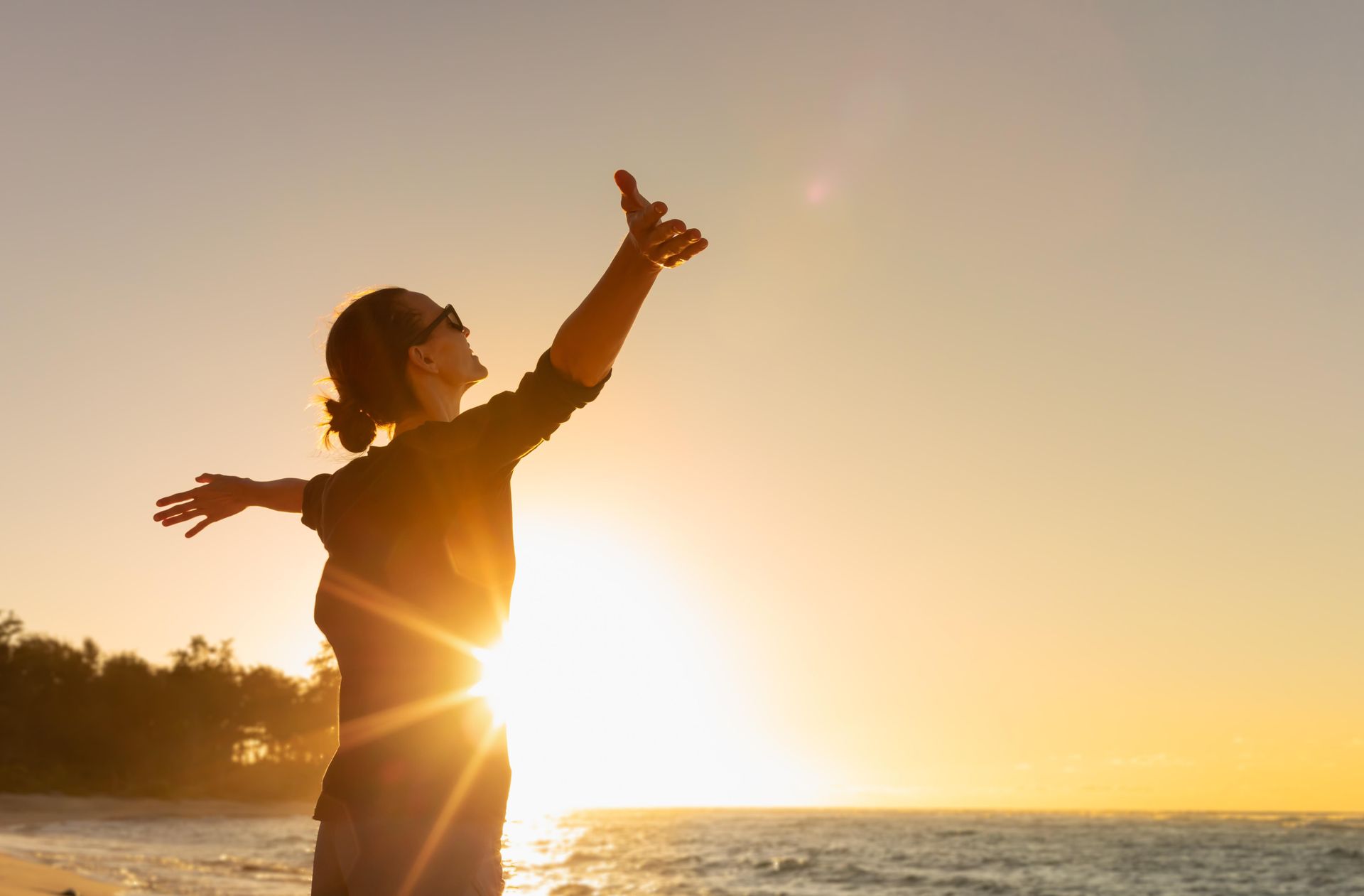 Person with arms raised towards bright sun on beach.