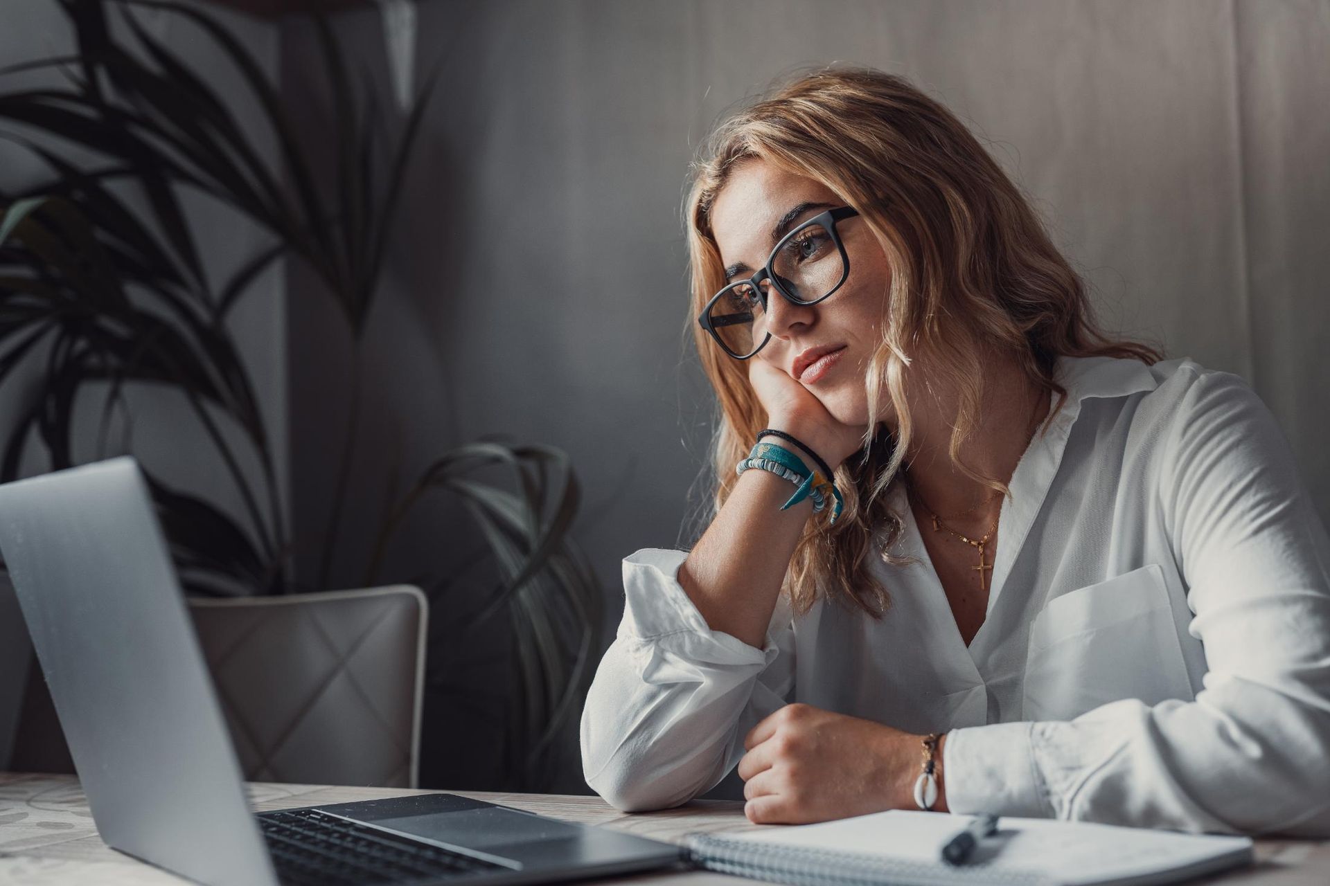 Woman wearing glasses, looking bored, leaning on hand while working on laptop.