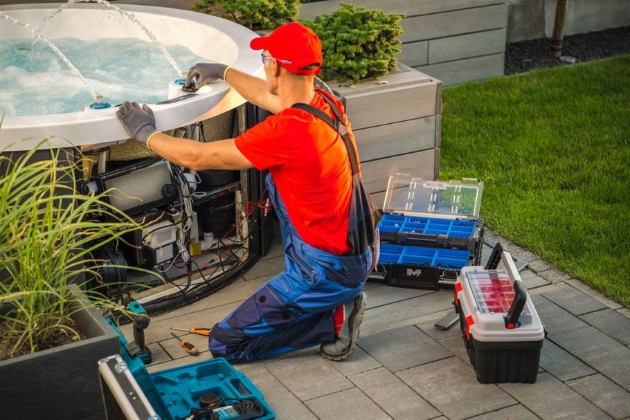 Technician in red cap and blue overalls repairing an outdoor hot tub beside tool cases.