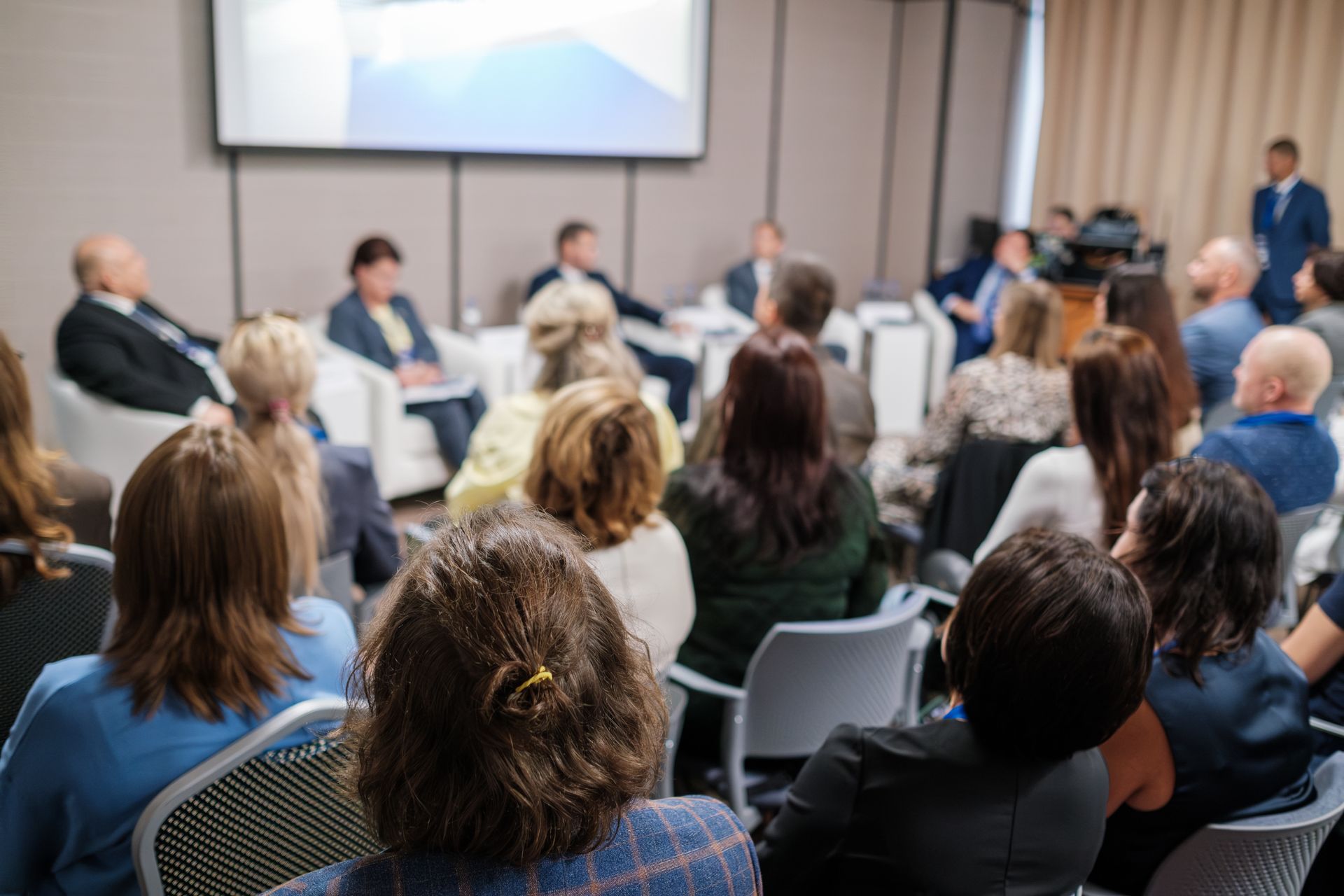 Conference audience seated before a panel discussion, with speakers at the front and a presentation screen above.