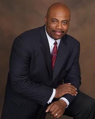 Man in a dark suit and red tie seated in a studio portrait, smiling at the camera.