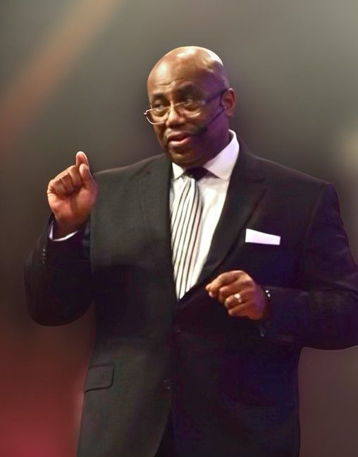 Man in a black suit gesturing onstage against a blurred dark background