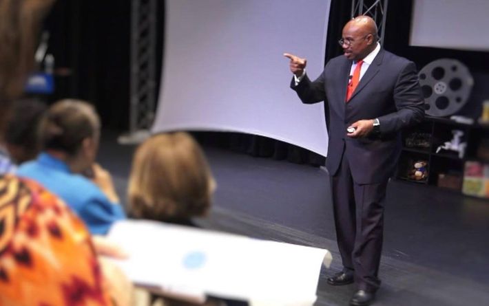 Speaker in dark suit and red tie gestures on stage before an audience.