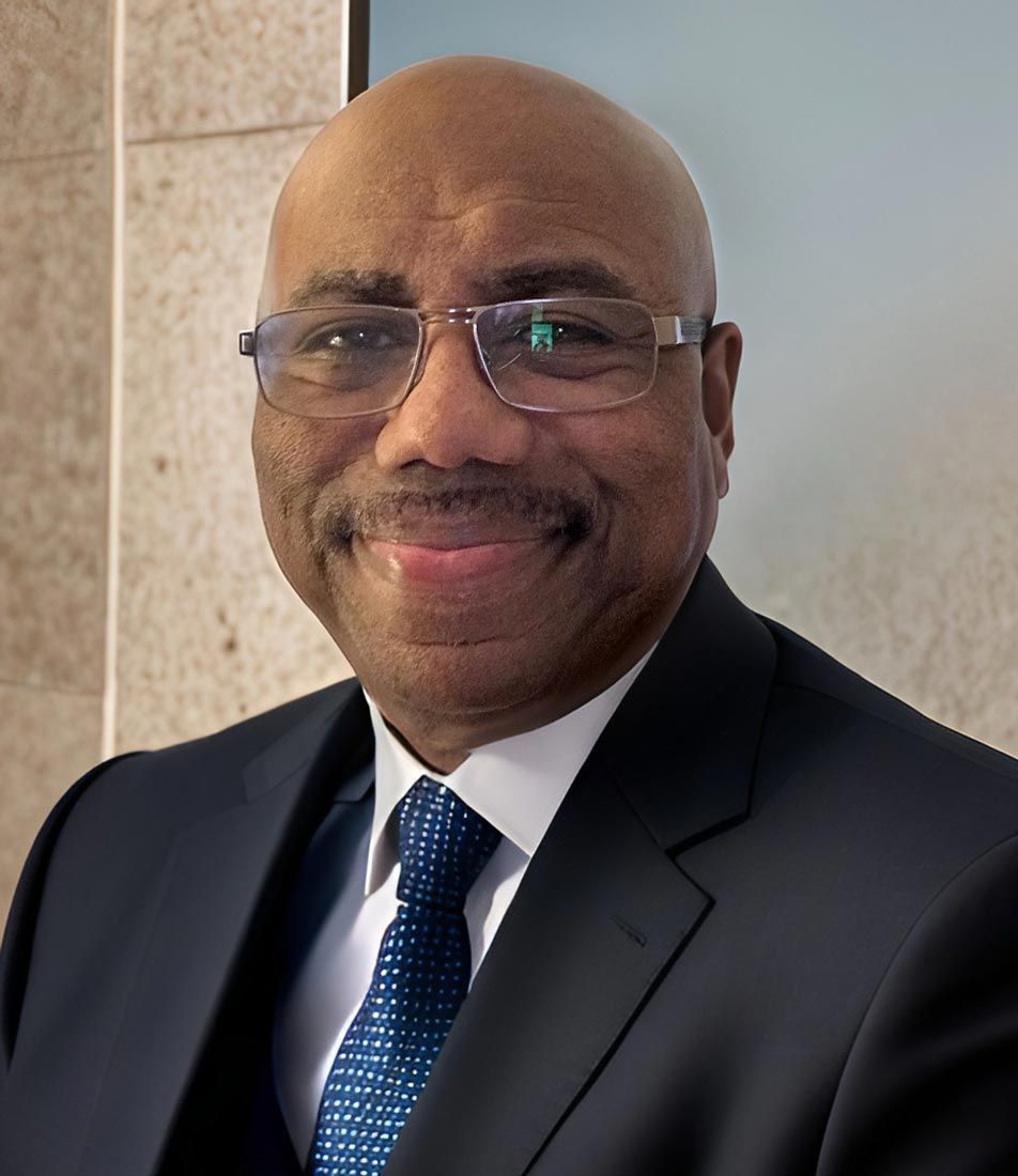 Smiling man in a dark suit and blue tie, wearing glasses, standing indoors against a tiled wall.