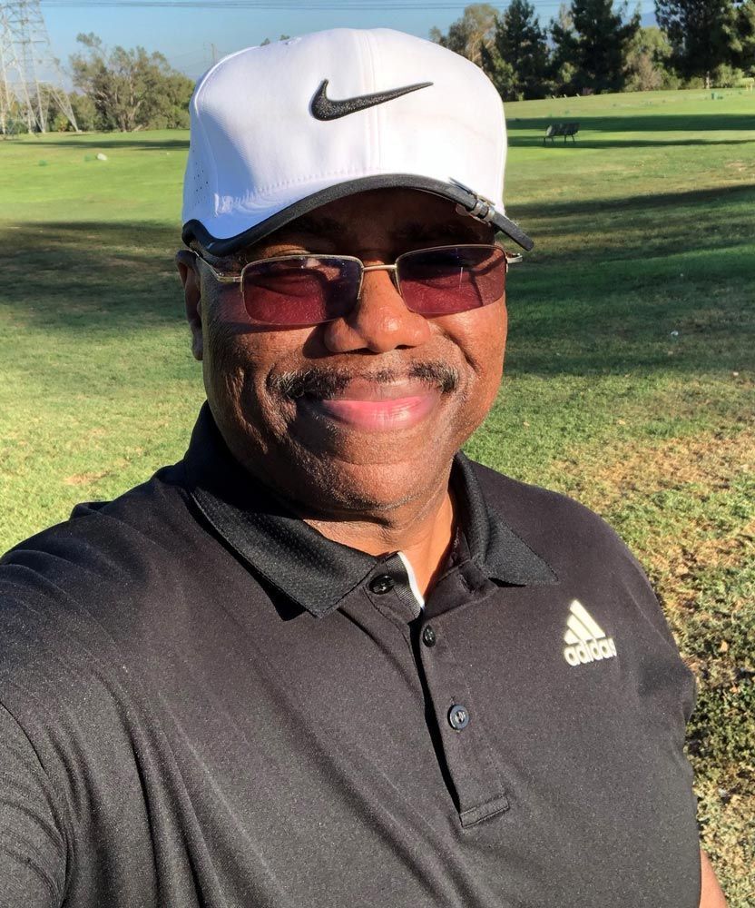 Smiling person in a white Nike cap and black polo shirt standing on a sunny golf course.