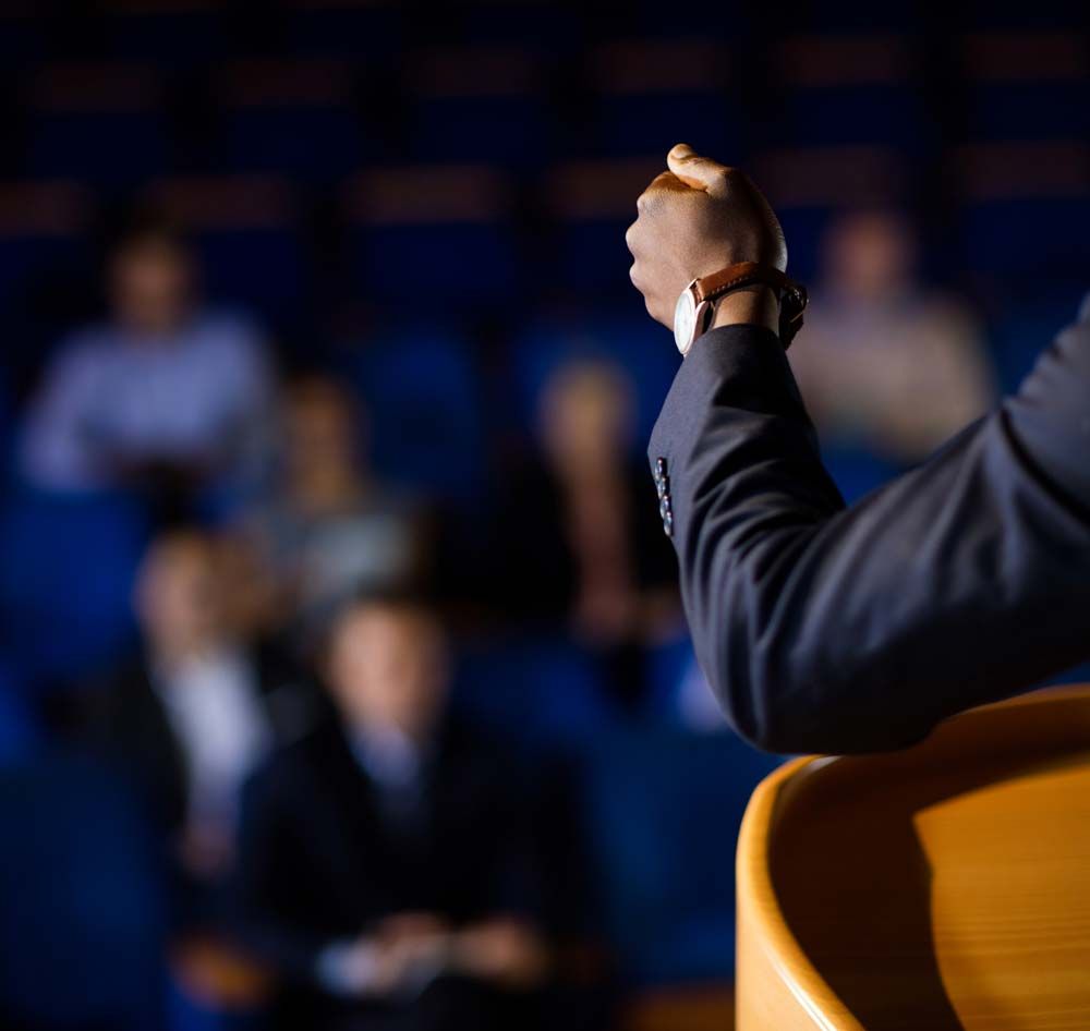 Speaker at podium addressing a blurred audience in a dim auditorium
