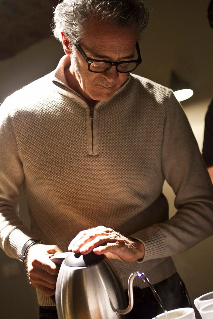 Man in a beige sweater pouring hot water from an electric kettle into a mug in a dimly lit kitchen