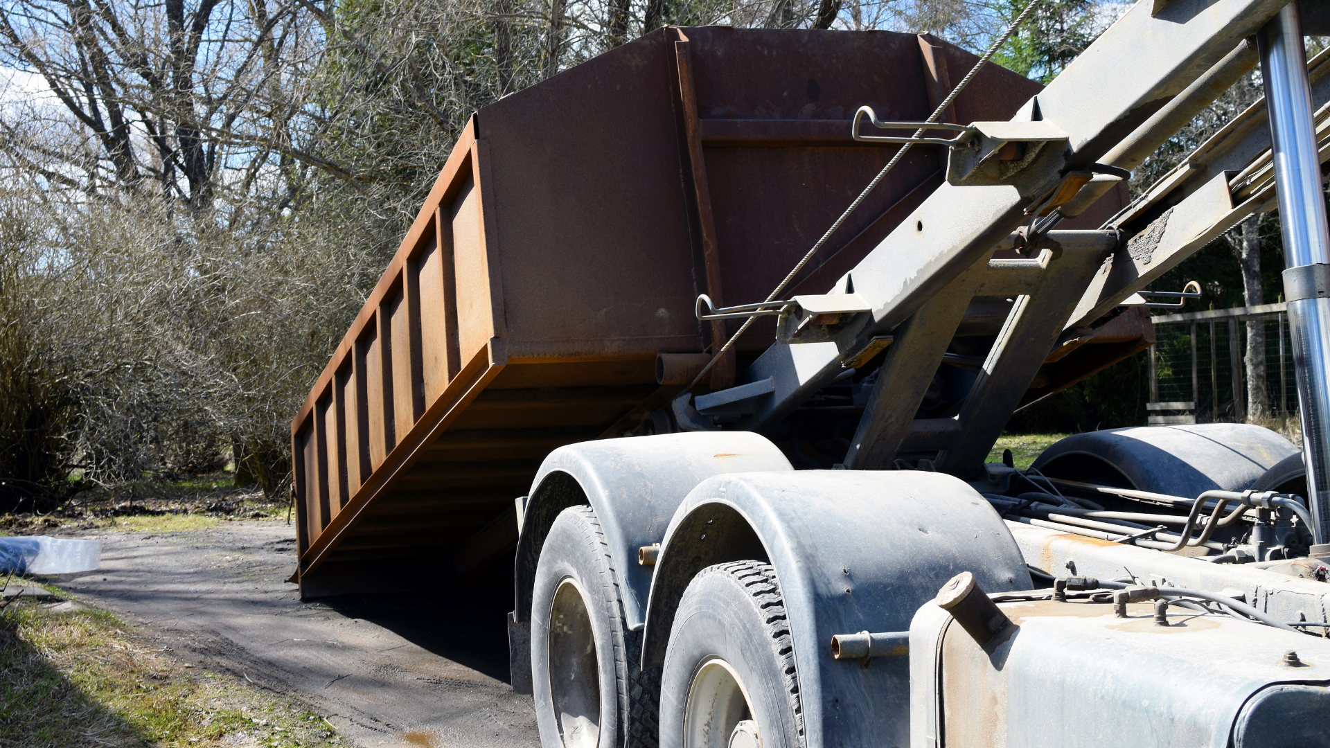 A dump truck is pulling a large brown dumpster down a dirt road.