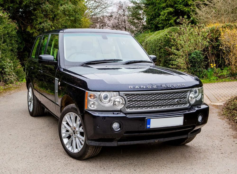 Black Range Rover parked on a paved road in front of green bushes and trees, sunny day.
