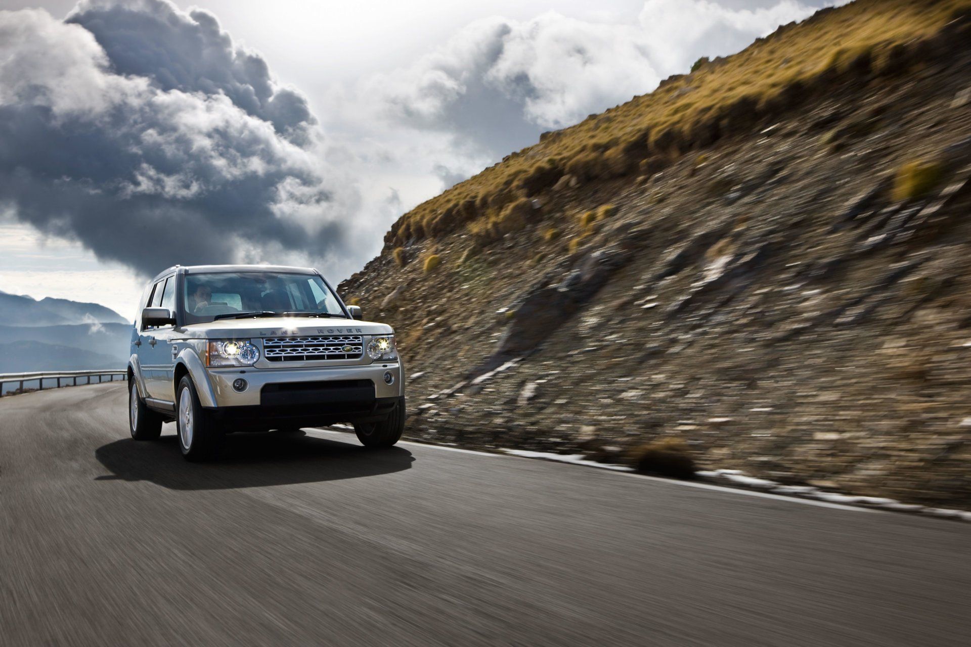 Silver SUV driving on a winding mountain road, with a cloudy sky and rocky hillside in the background.