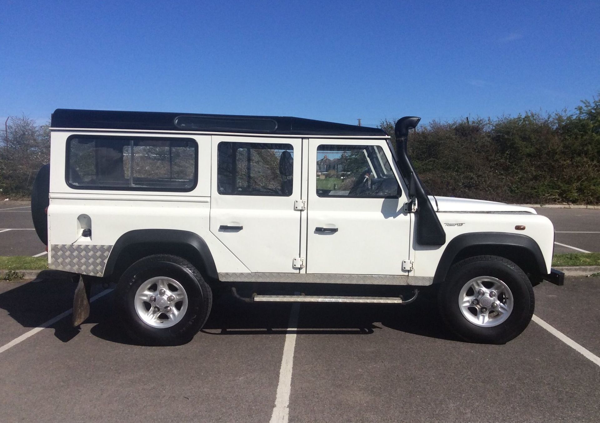 White Land Rover Defender parked in a paved lot with a blue sky background. It has a black roof and snorkel.