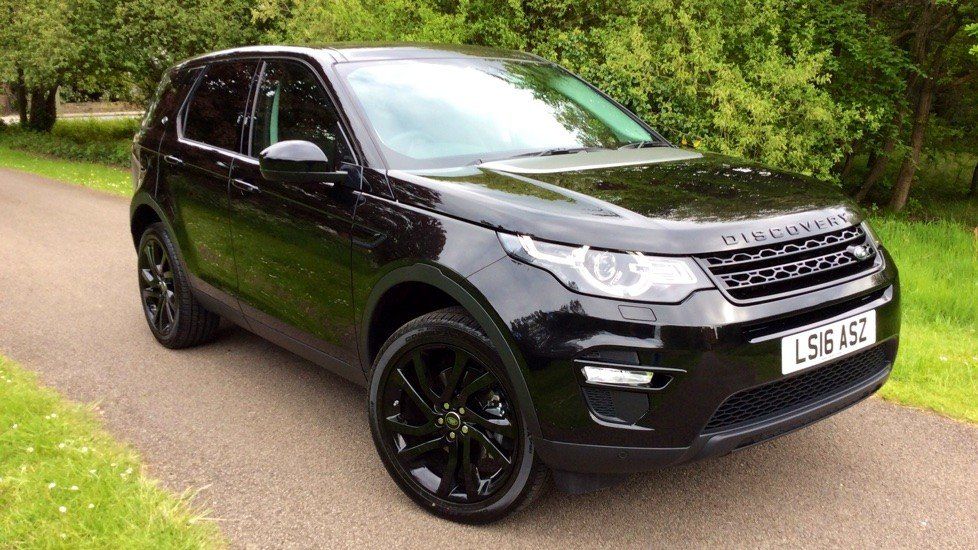 Black Land Rover Discovery Sport parked on a paved road, with black rims, against a green background.