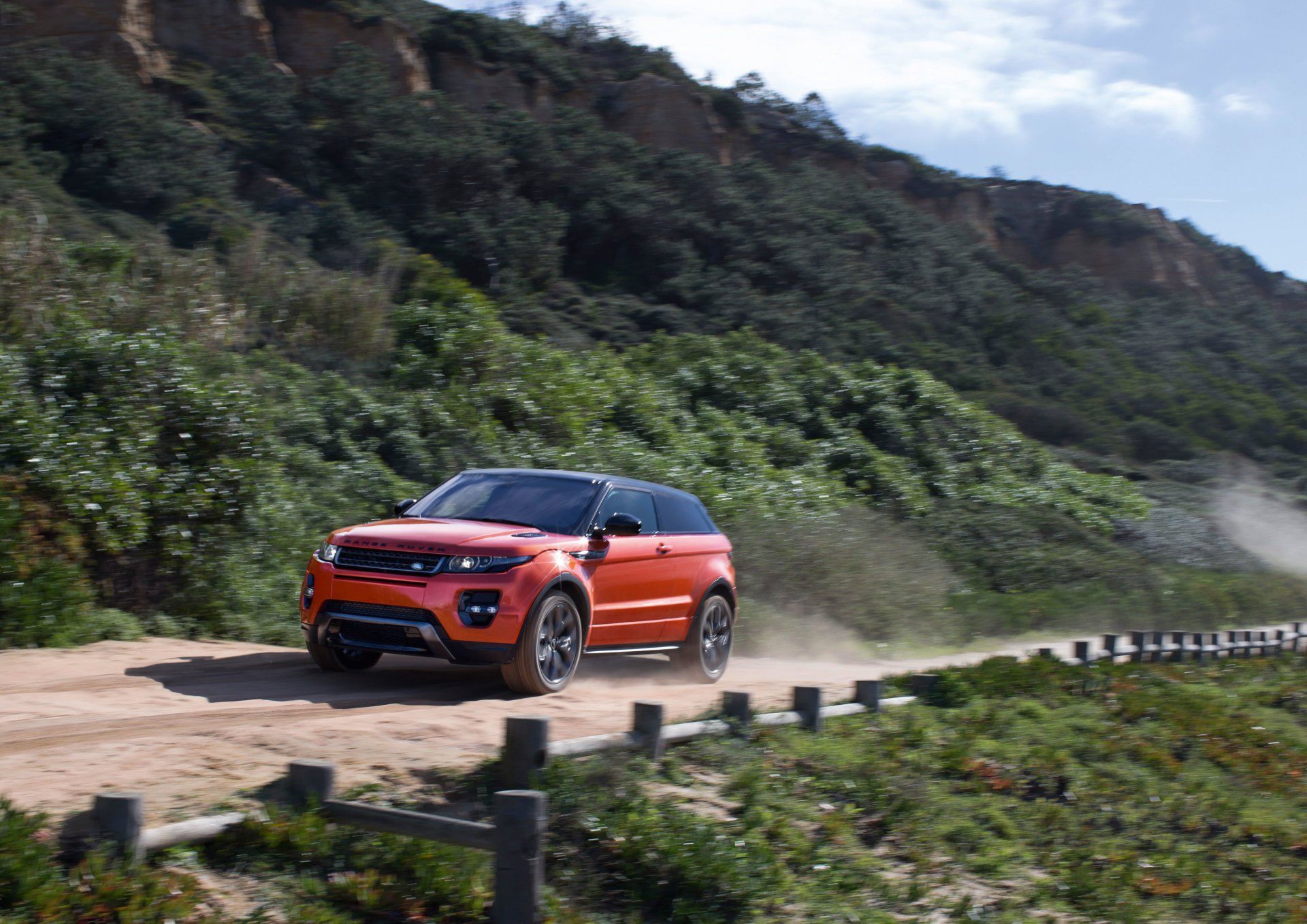 Orange Range Rover SUV driving on a dirt road along a coastal cliffside, kicking up dust.